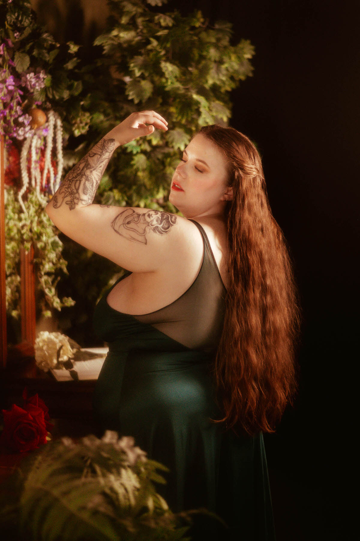 A woman in a sheer-back teal gown lifts her arm in a graceful pose, surrounded by trailing florals and greenery in a fine art pre-Raphaelite boudoir session at a Dallas boudoir studio.