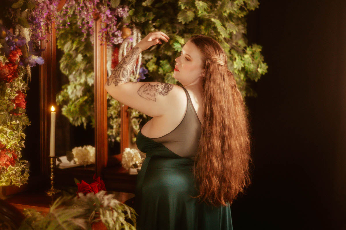 A woman in a sheer-back teal gown poses with her arm lifted in front of a candlelit mirror framed with florals and greenery during a pre-Raphaelite boudoir session at a Dallas boudoir studio.