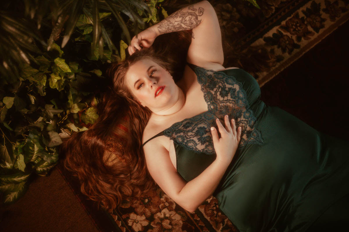 A curvy woman in a teal lace gown lies on a vintage rug beside lush greenery, gazing upward with her hand in her hair during a fine art pre-Raphaelite boudoir session at a Dallas boudoir studio.
