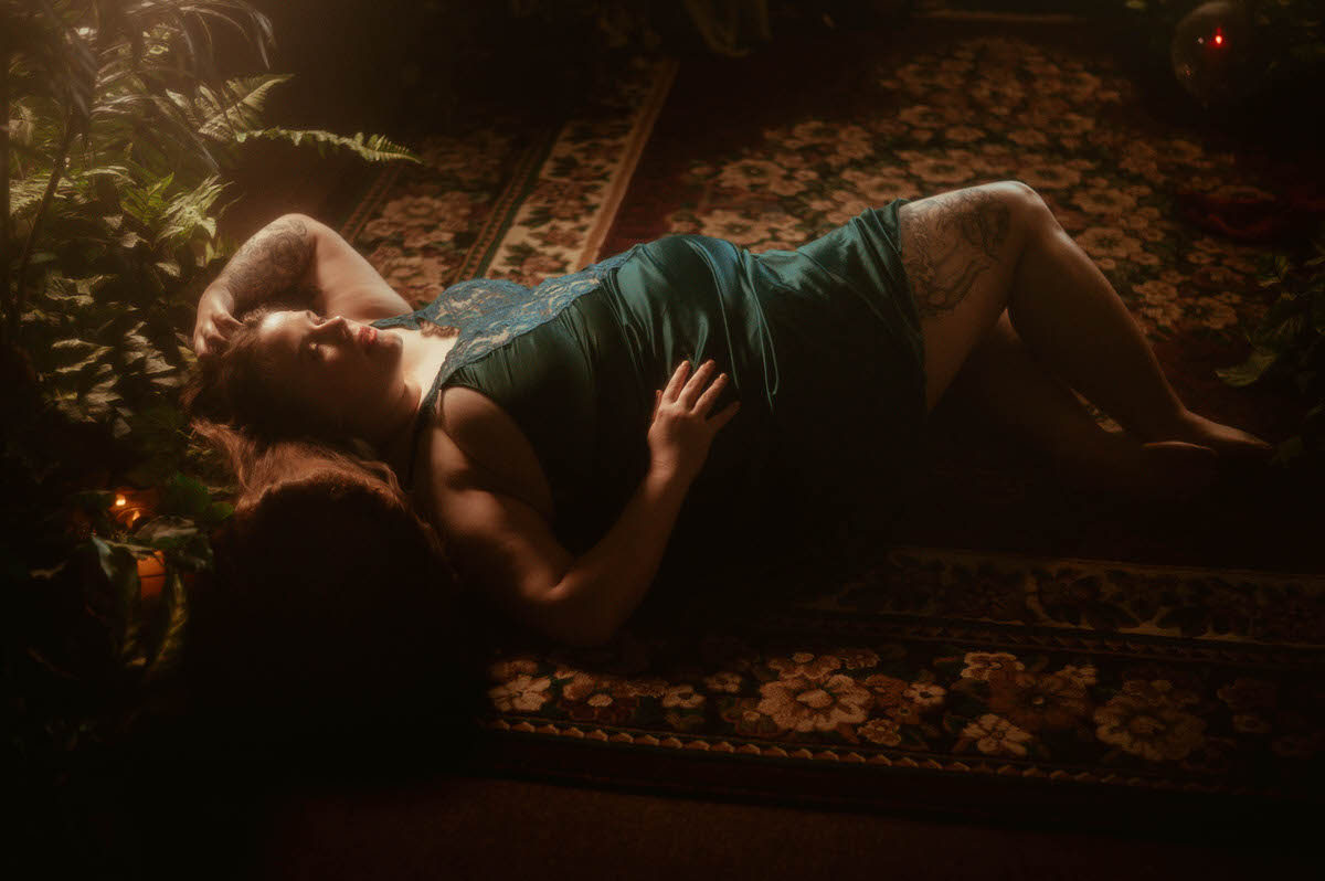 A woman in a teal lace gown lies on a floral vintage rug, surrounded by ivy and candlelight, styled in a lush pre-Raphaelite boudoir session at a Dallas boudoir studio.