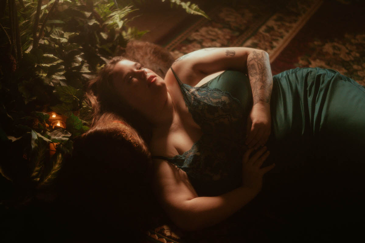 A woman in a teal lace nightgown lies in shadowy candlelight beside ivy-covered floors and vintage rugs, styled in a moody pre-Raphaelite boudoir session at a Dallas boudoir studio.