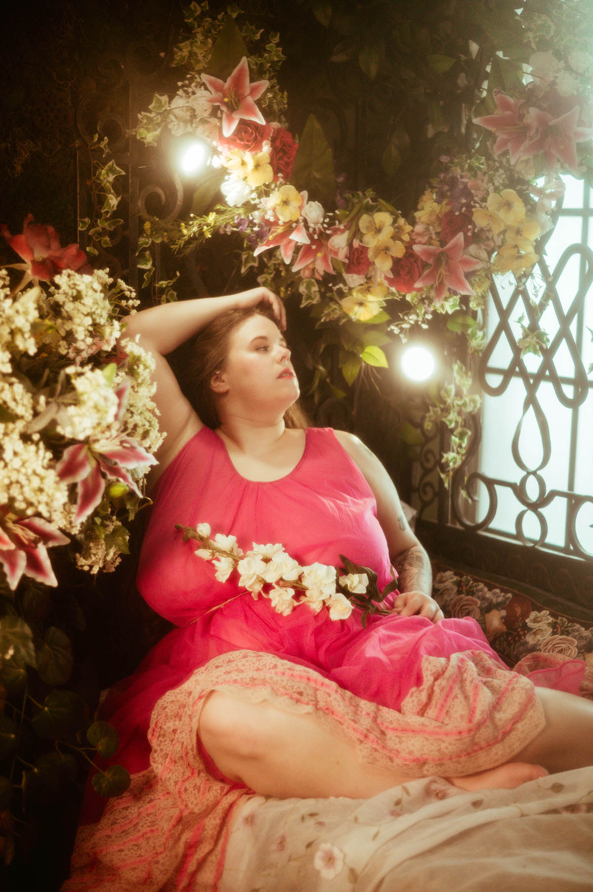 A woman in a bright pink vintage gown reclines among cascading floral arrangements and soft lighting, channeling a dreamy pre-Raphaelite boudoir aesthetic at a Dallas boudoir studio.