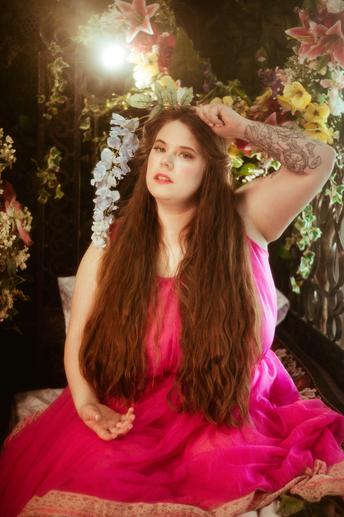 A woman in a vivid pink vintage gown poses with cascading blue flowers in her hair, surrounded by blooms and soft light during a whimsical pre-Raphaelite boudoir session at a Dallas boudoir studio.