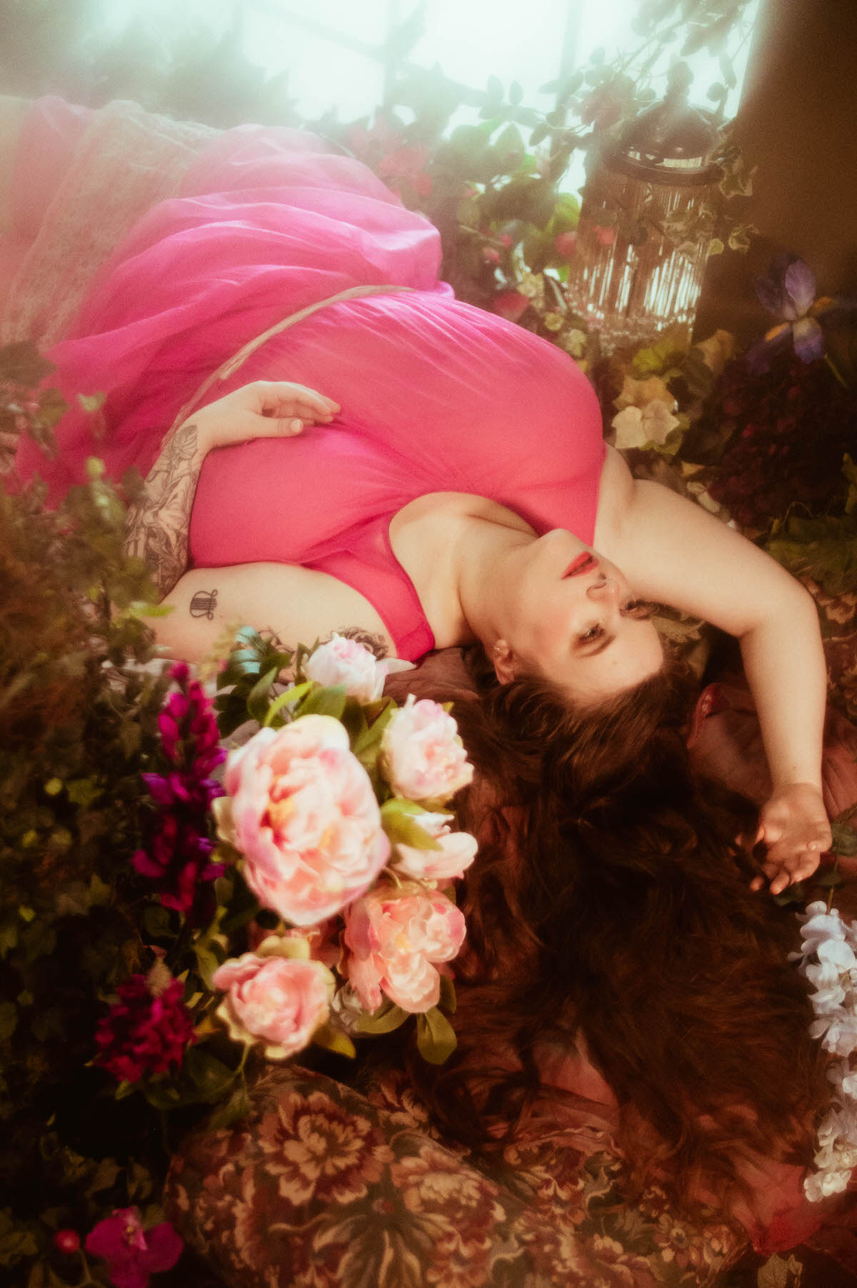 Reclining on a bed of floral textiles and surrounded by pink peonies, a woman in a flowing magenta dress gazes dreamily upward in this romantic pre-Raphaelite boudoir portrait.