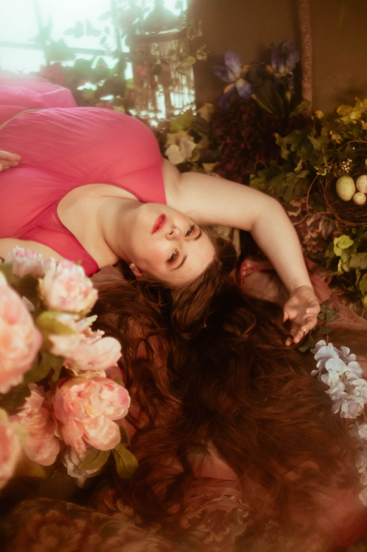 A woman in a flowing pink dress lies on a floral-covered bed, framed by pastel roses and soft greenery, during a dreamy pre-Raphaelite boudoir photoshoot in Dallas.