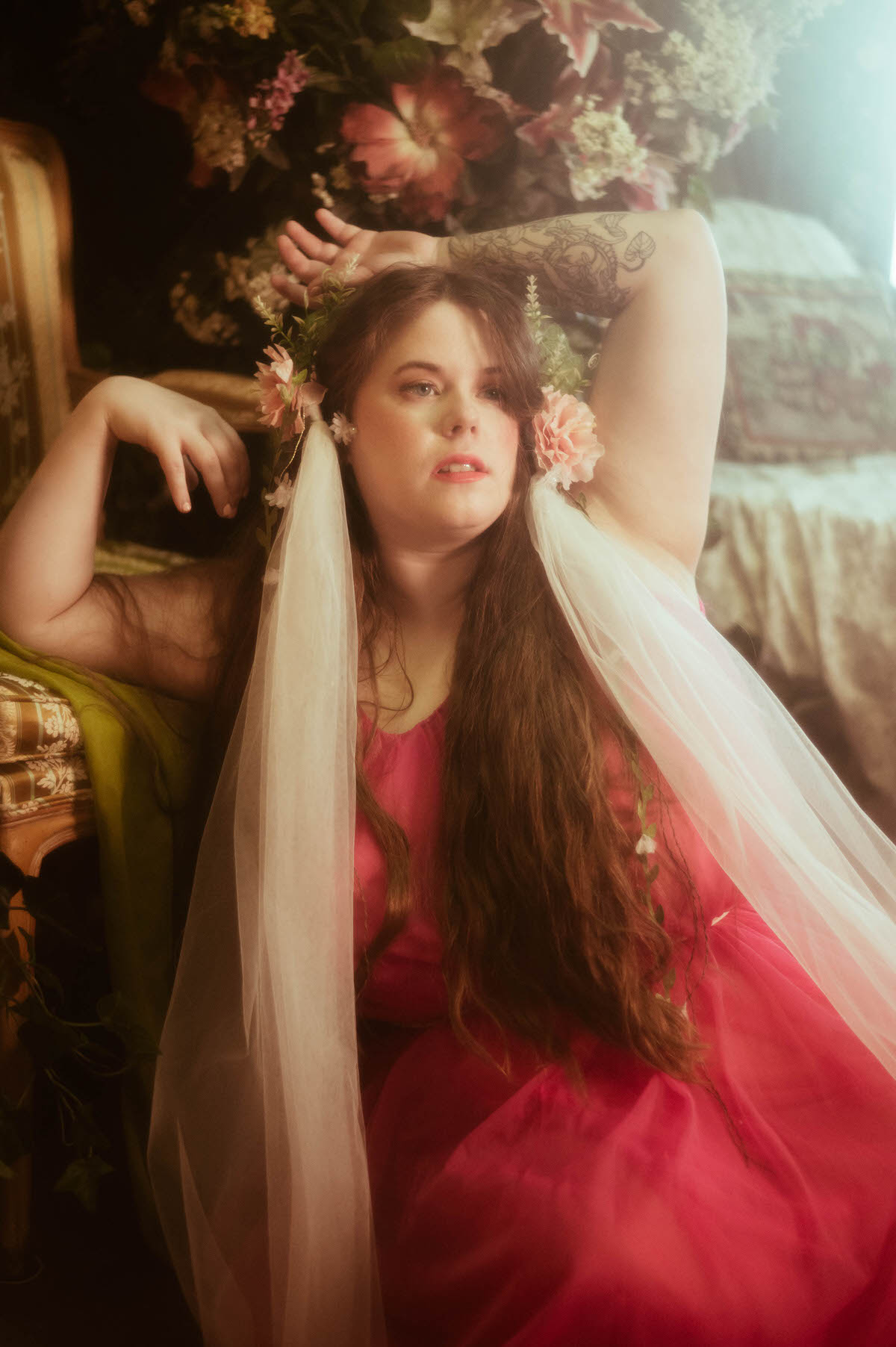 In a romantic pre-Raphaelite boudoir scene, a woman in a pink gown reclines beside a vintage chair, wearing a floral crown with trailing white tulle and gazing wistfully into soft window light.