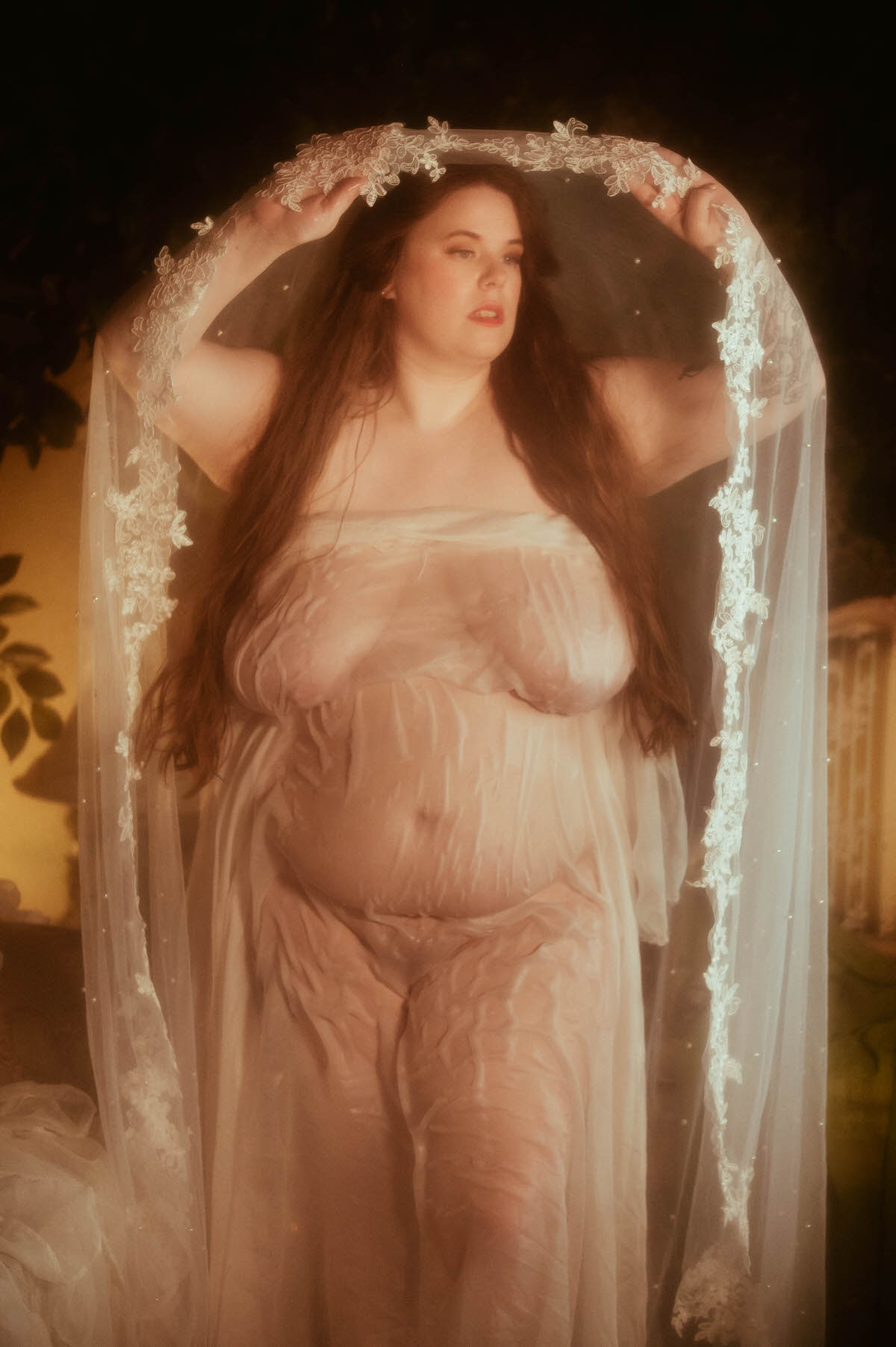 A nude plus-sized woman draped in sheer fabric stands gracefully beneath a pearl-studded veil, channeling pre-Raphaelite softness and artistic boudoir elegance in warm golden light.