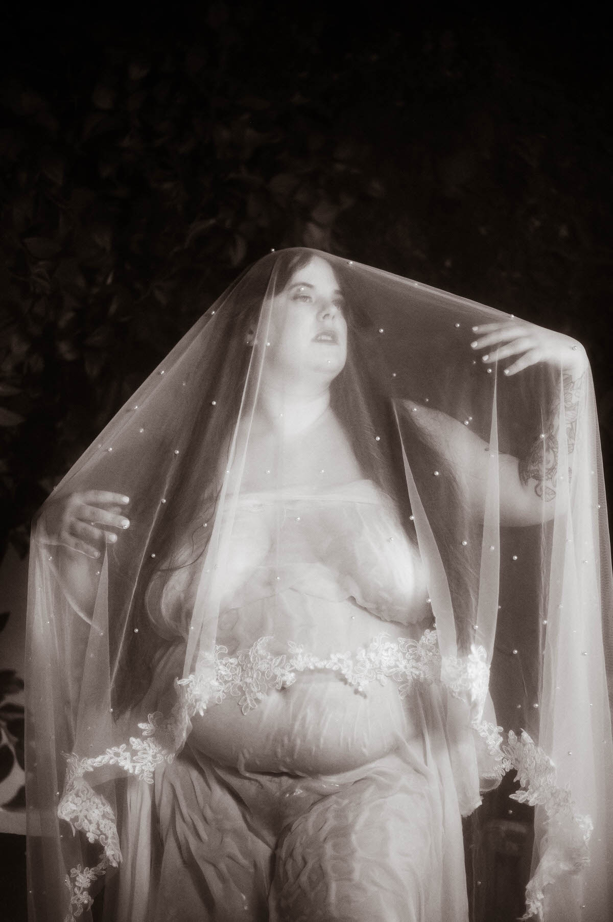 Black and white portrait of a nude curvy woman veiled in sheer fabric with pearl accents and lace trim, her ethereal pose channeling pre-Raphaelite elegance and fantasy boudoir style.