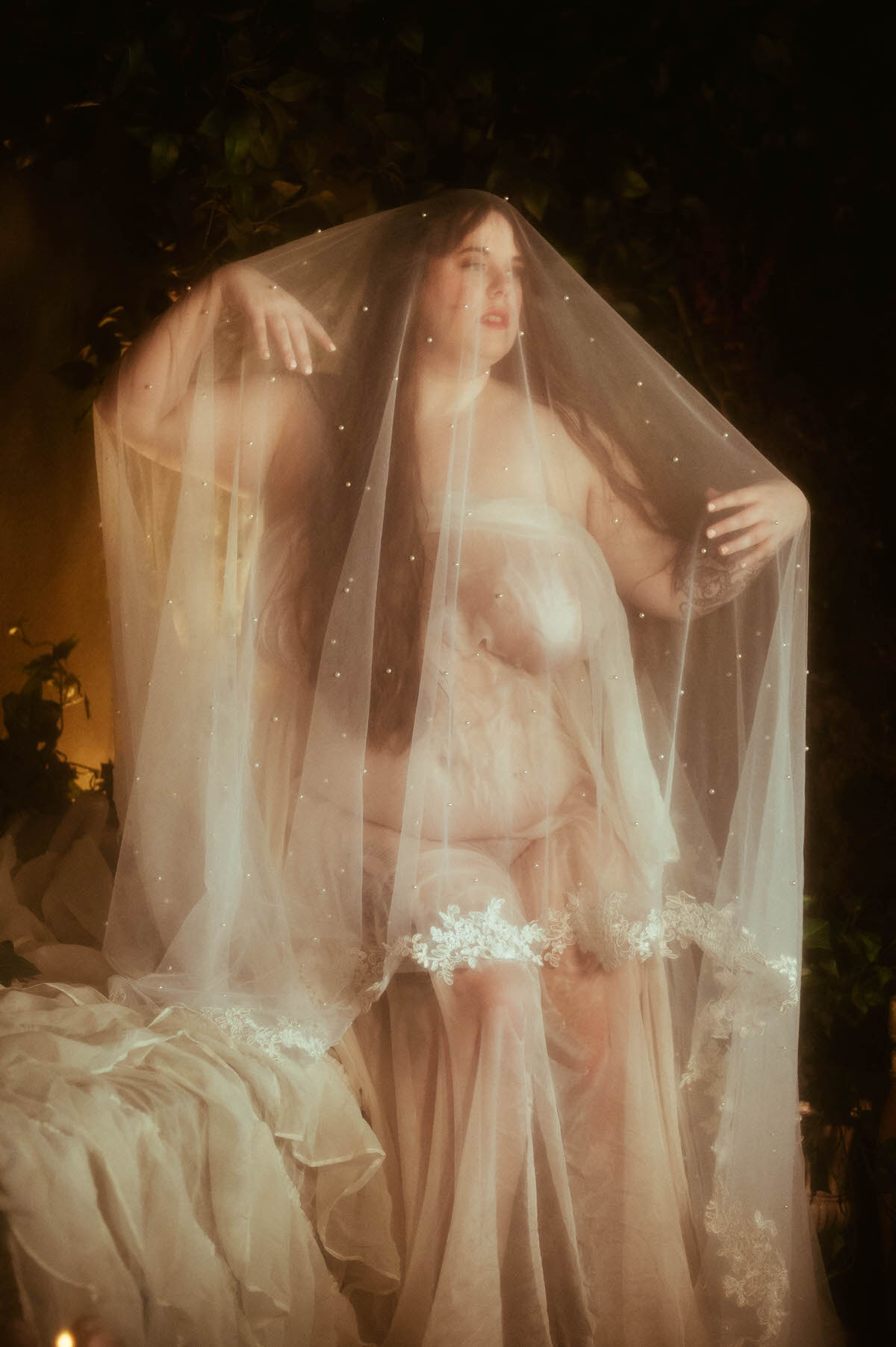 A nude curvy woman draped in sheer white fabric and a pearl-dotted veil poses ethereally on a bed, evoking a dreamy pre-Raphaelite boudoir aesthetic surrounded by soft shadows.