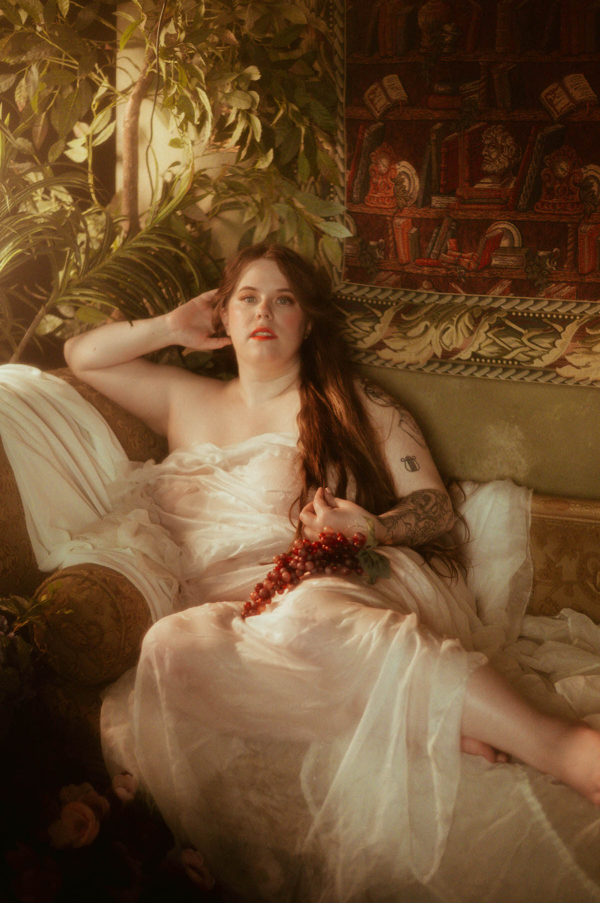 A woman reclines on a vintage settee draped in white fabric, holding a bunch of grapes, evoking Pre-Raphaelite opulence with rich textures, greenery, and classical tapestry.