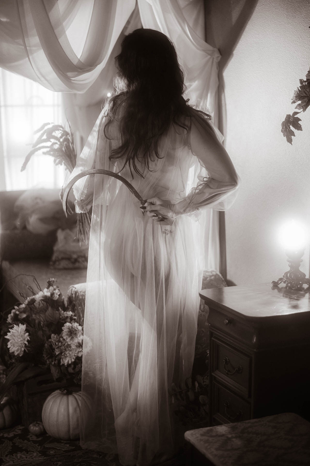 A moody fall photos black and white silhouette captures a woman from behind in sheer fabric holding a sickle and dried botanicals before a sunlit window, inspired by the painting Allegory of Summer. Royal Lune Photo.
