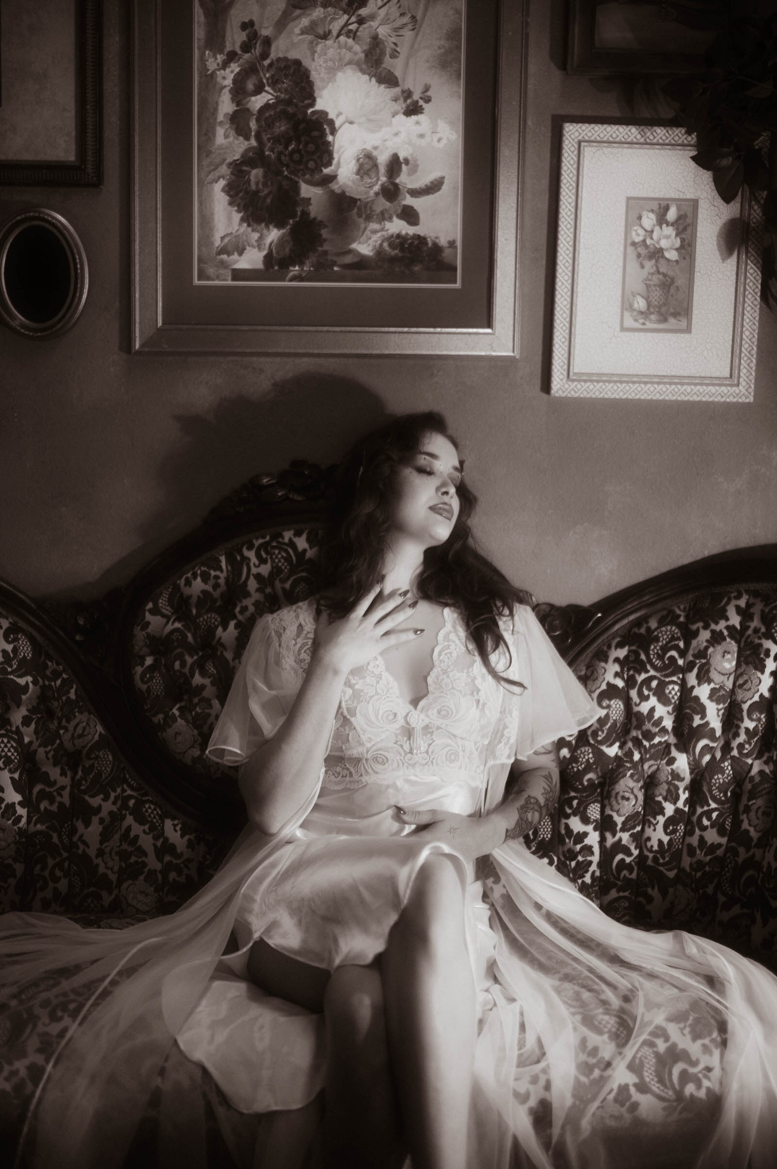 This moody fall photos black and white portrait features a woman in sheer lace lounging against an antique tufted sofa with her head tilted back in soft repose. Royal Lune Photo.