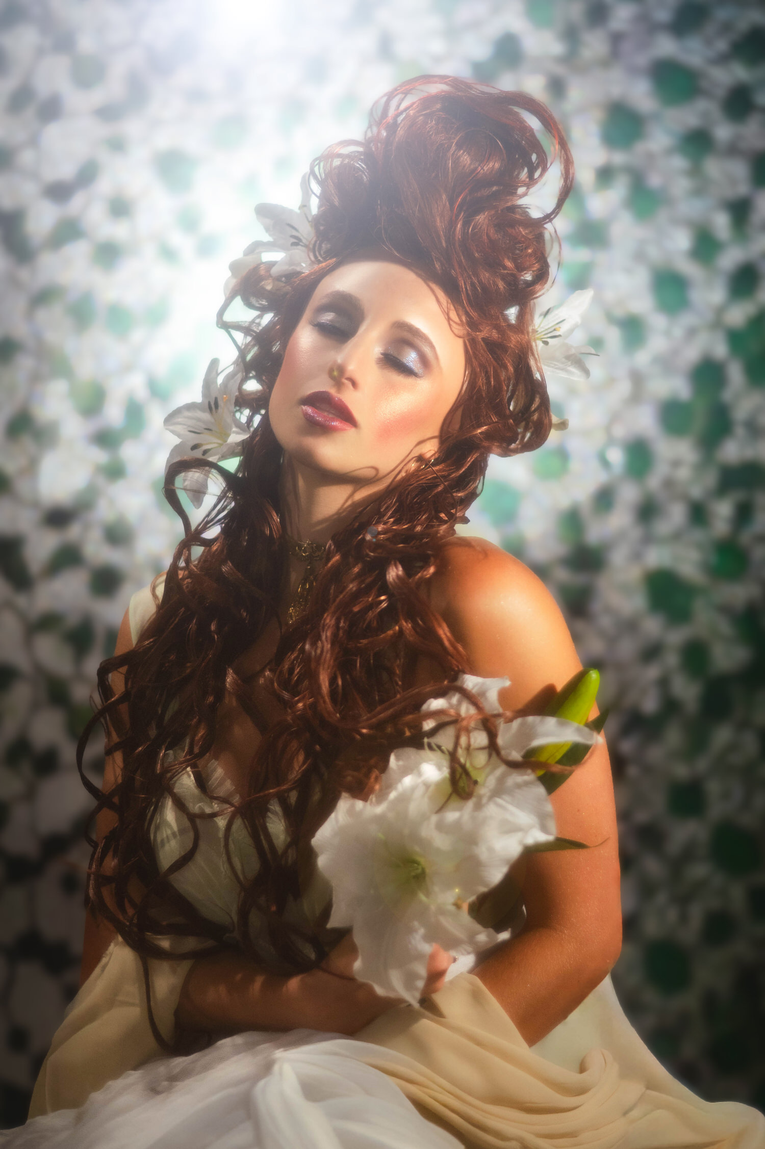 Fine art Art Nouveau photoshoot. A seated woman in a sheer sage dress framed by a backlit leafy background window, holding white floral stems.