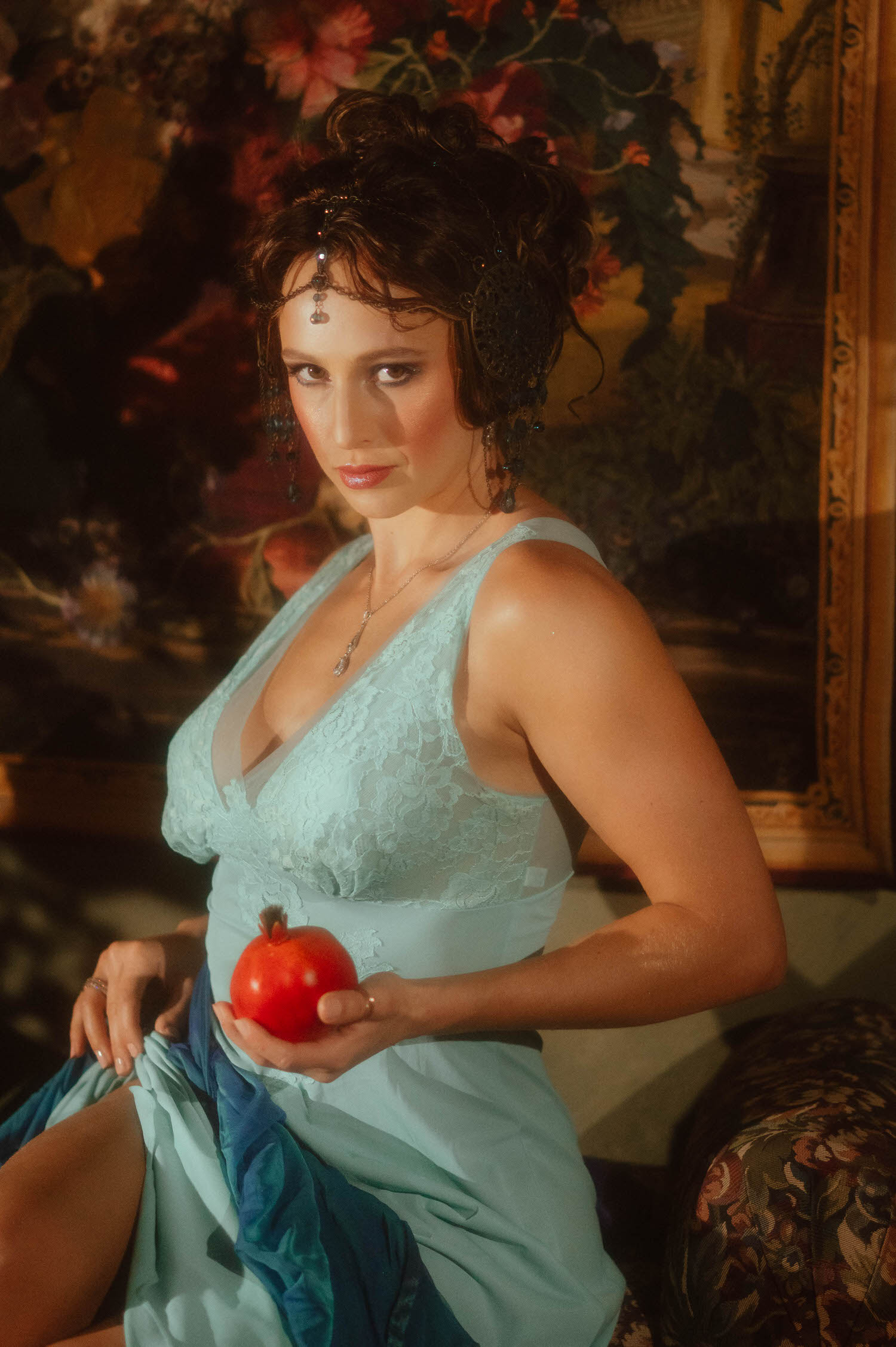 A fine art portrait of a woman in a lace blue gown holding a red pomegranate. Classic Alphonse Mucha styling inside a Dallas area photography studio.