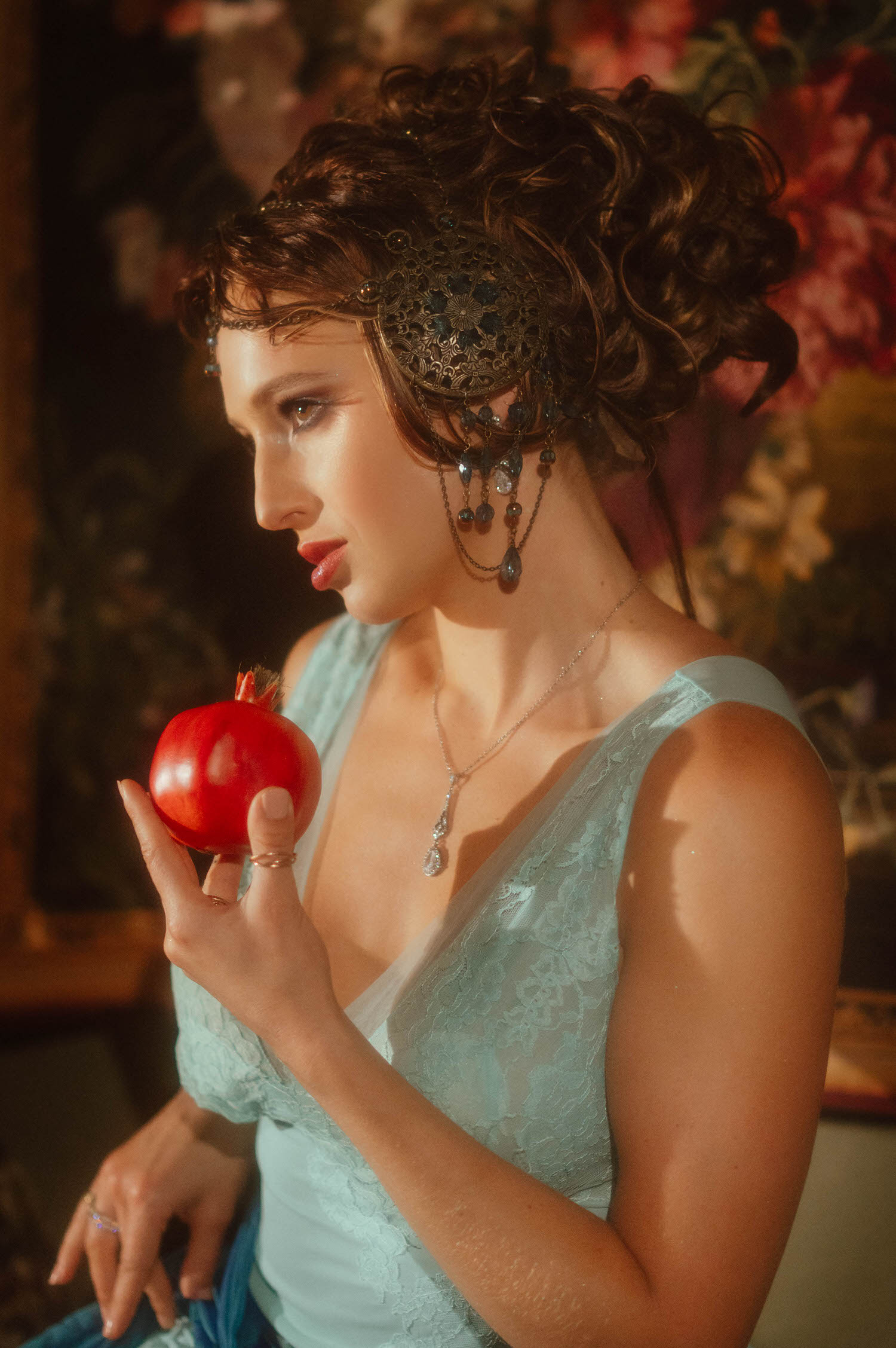 Art Nouveau style portrait highlighting an ornate metal headpiece and a blue dress. The model looks down while holding a pomegranate at Royal Lune Studio.