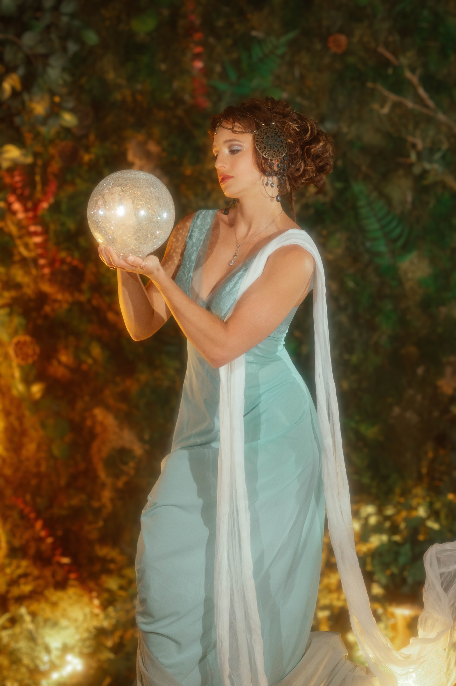 Fine art portrait inside Royal Lune Studio near Dallas. A model in a flowing dress looks down thoughtfully at a luminous orb against a background of green ivy.
