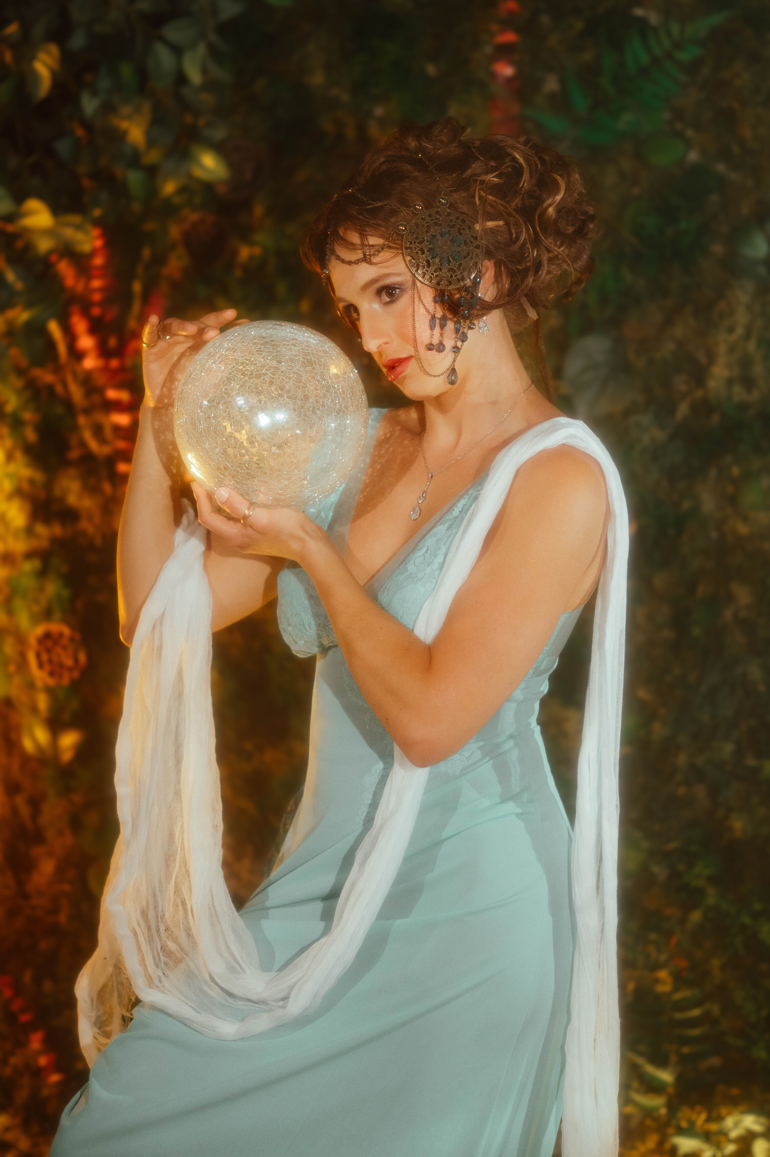 Fine art portrait showcasing classic Art Nouveau style. A model at Royal Lune Studio holds a luminous crystal orb prop against a leafy green background.