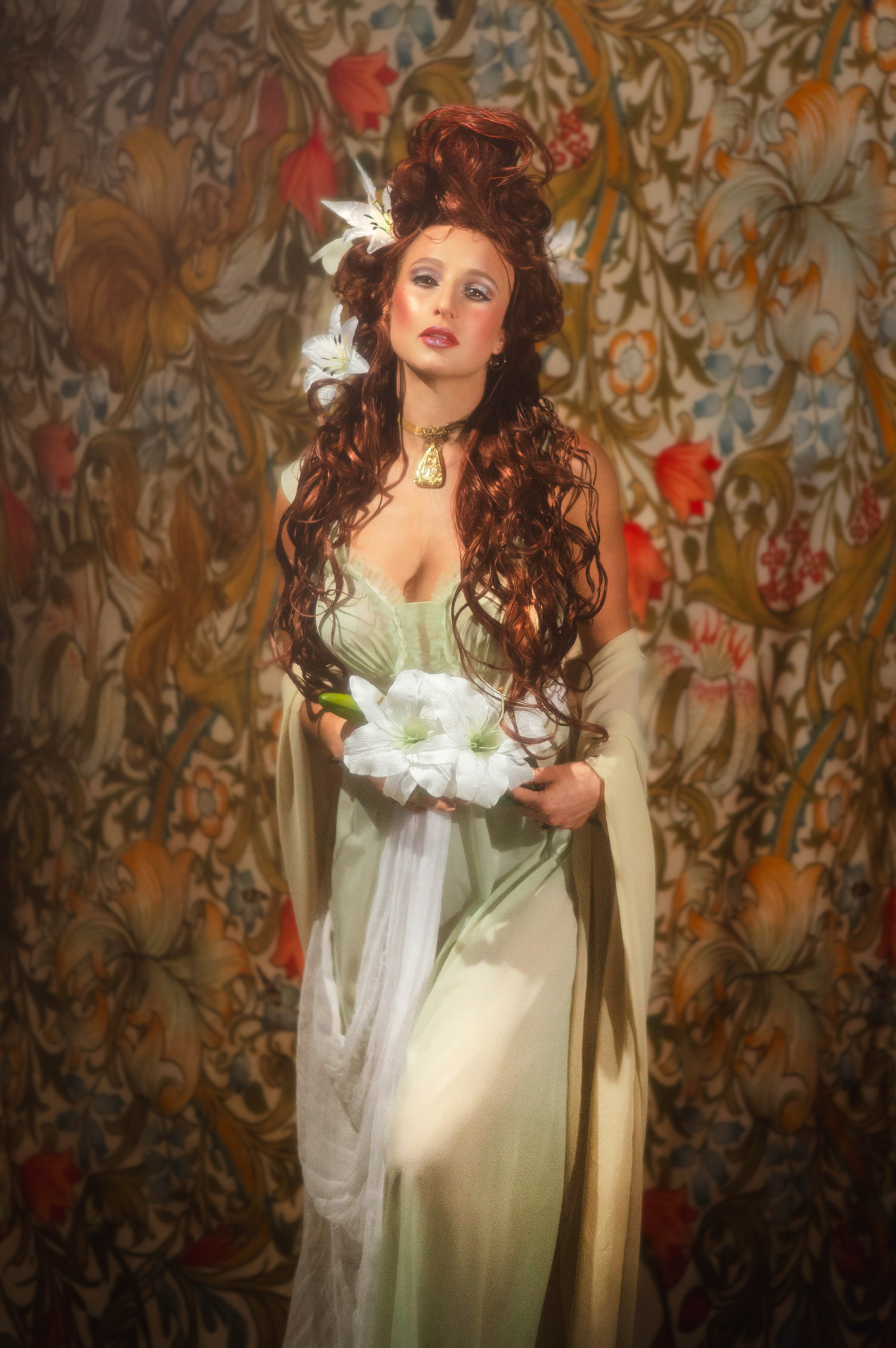 Alphonse Mucha inspired Art Nouveau photoshoot. Full-length portrait of a model in a pale green vintage gown holding lilies against a detailed vintage floral pattern.