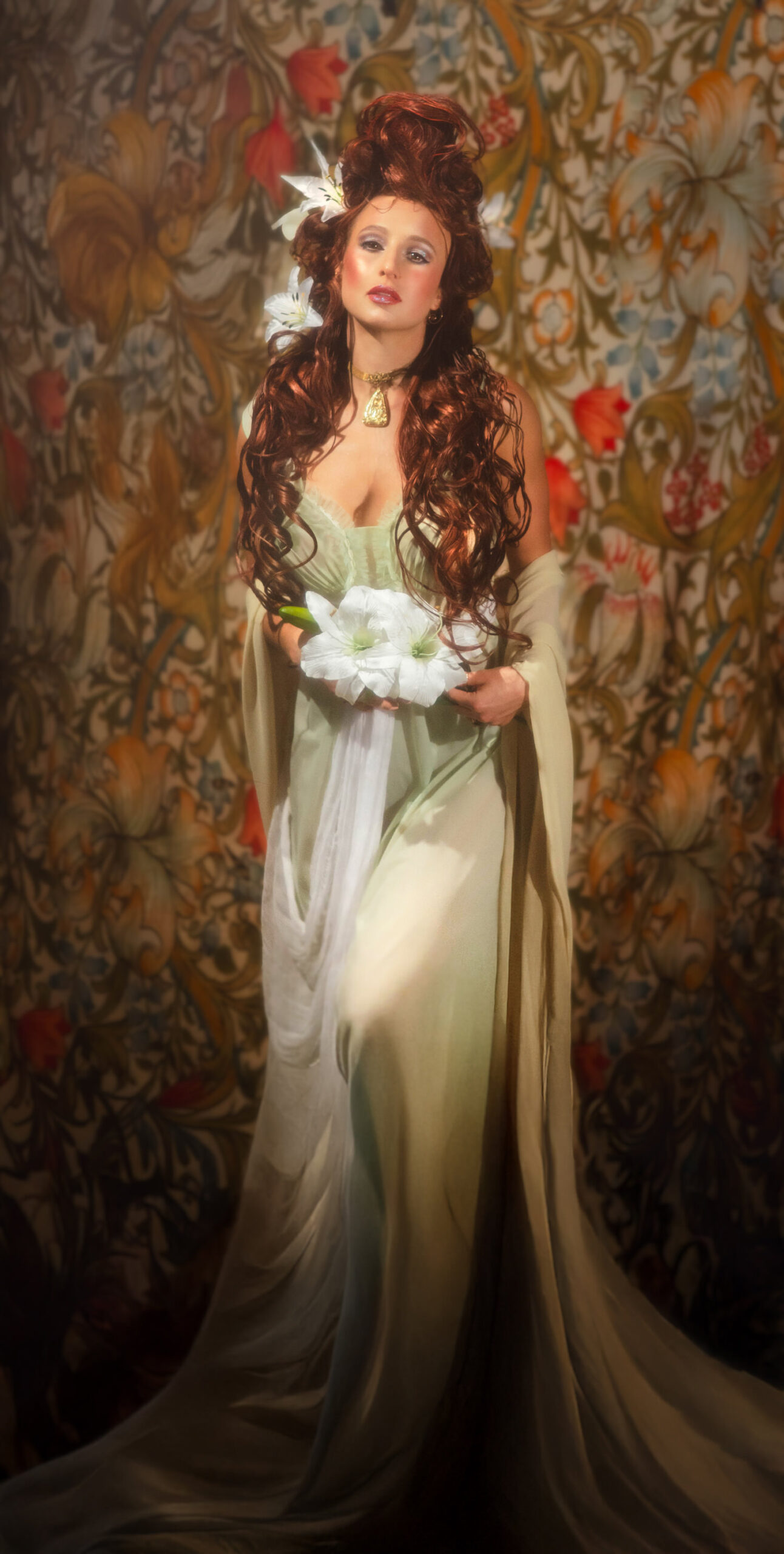 Art Nouveau photoshoot featuring a model with long red hair and white lilies, posing in a green gown against a floral tapestry background. Alphonse Mucha portrait style.