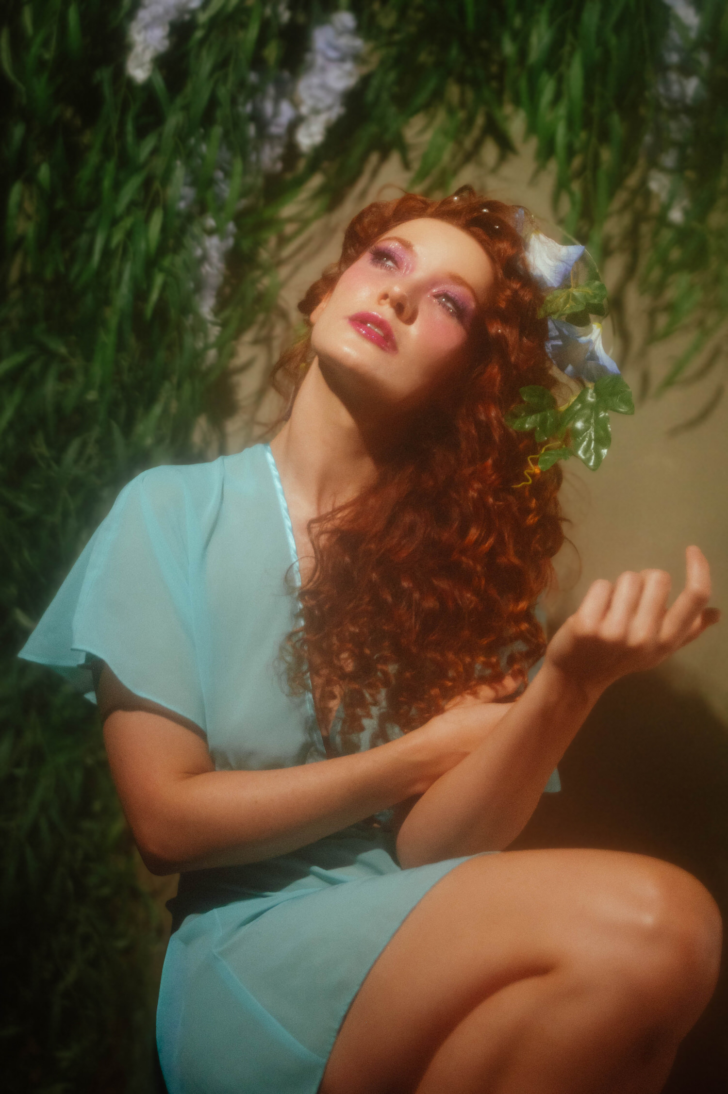 A red haired model in a vintage teal nightgown looks upward beneath hanging greenery, captured for a fine art Midsummer Night's Dream photoshoot. This dreamy photography studio session took place near Dallas. Royal Lune Photo.