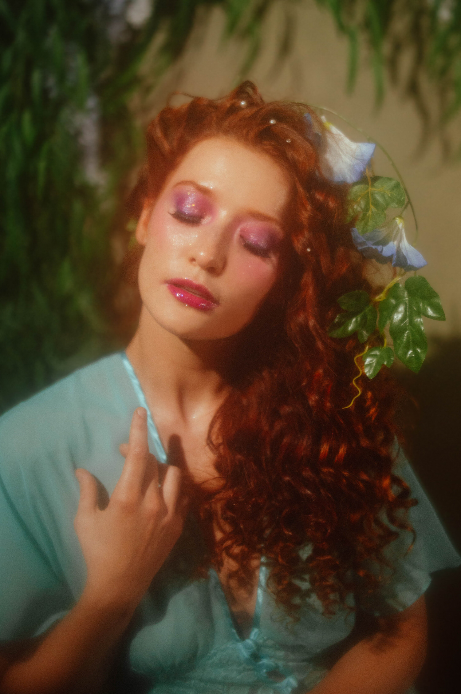 A close up portrait of a model with curly red hair and flowers tucked into her waves, captured in a dreamy photography style for a Midsummer Night's Dream inspired session. This fine art image was created at a Dallas area studio in a vintage teal nightgown. Royal Lune Photo.
