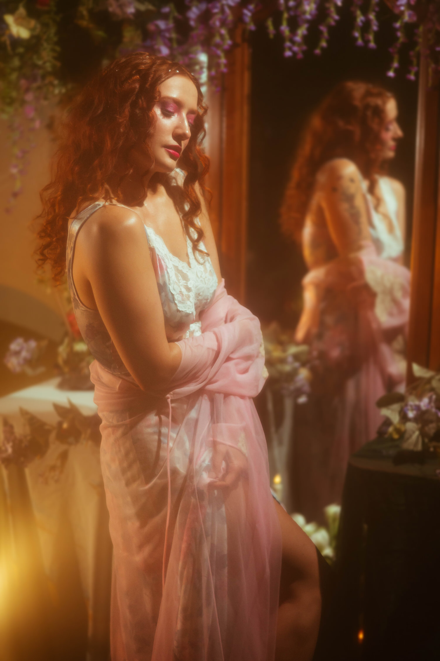 A woman in a vintage lace nightgown and flowing pink robe stands in front of an antique mirror surrounded by purple florals, reflecting the dreamy photography style of this Dallas area studio session. The soft warm lighting enhances the fairytale mood of the Midsummer Night's Dream theme. Royal Lune Photo.