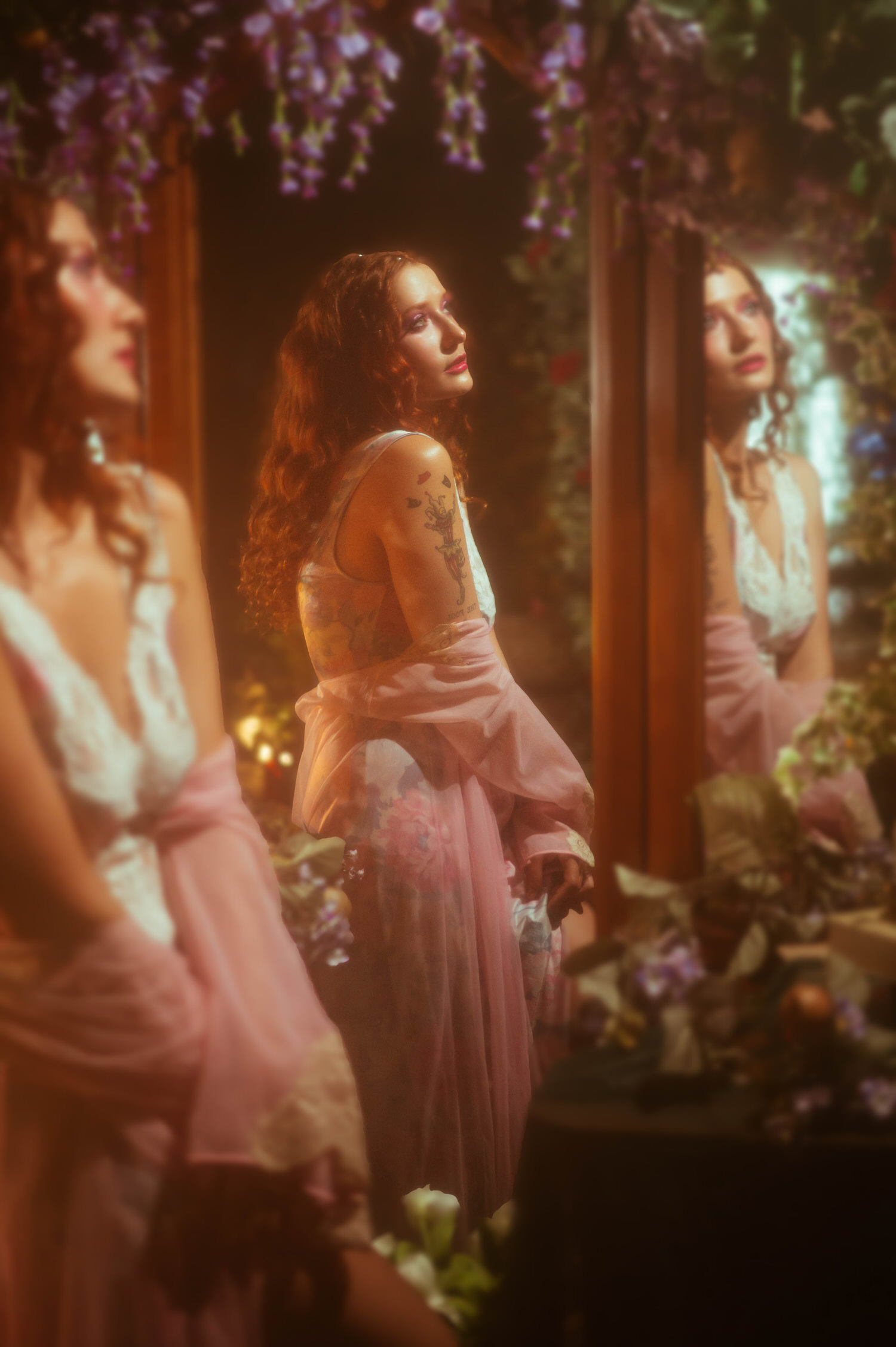 A red haired model in a vintage nightgown and sheer pink robe stands before a mirrored reflection, surrounded by hanging florals in a dreamy photography studio inspired by A Midsummer Night's Dream. The layered vintage wardrobe and warm tones capture the fine art aesthetic of this Dallas photography session. Royal Lune Photo.