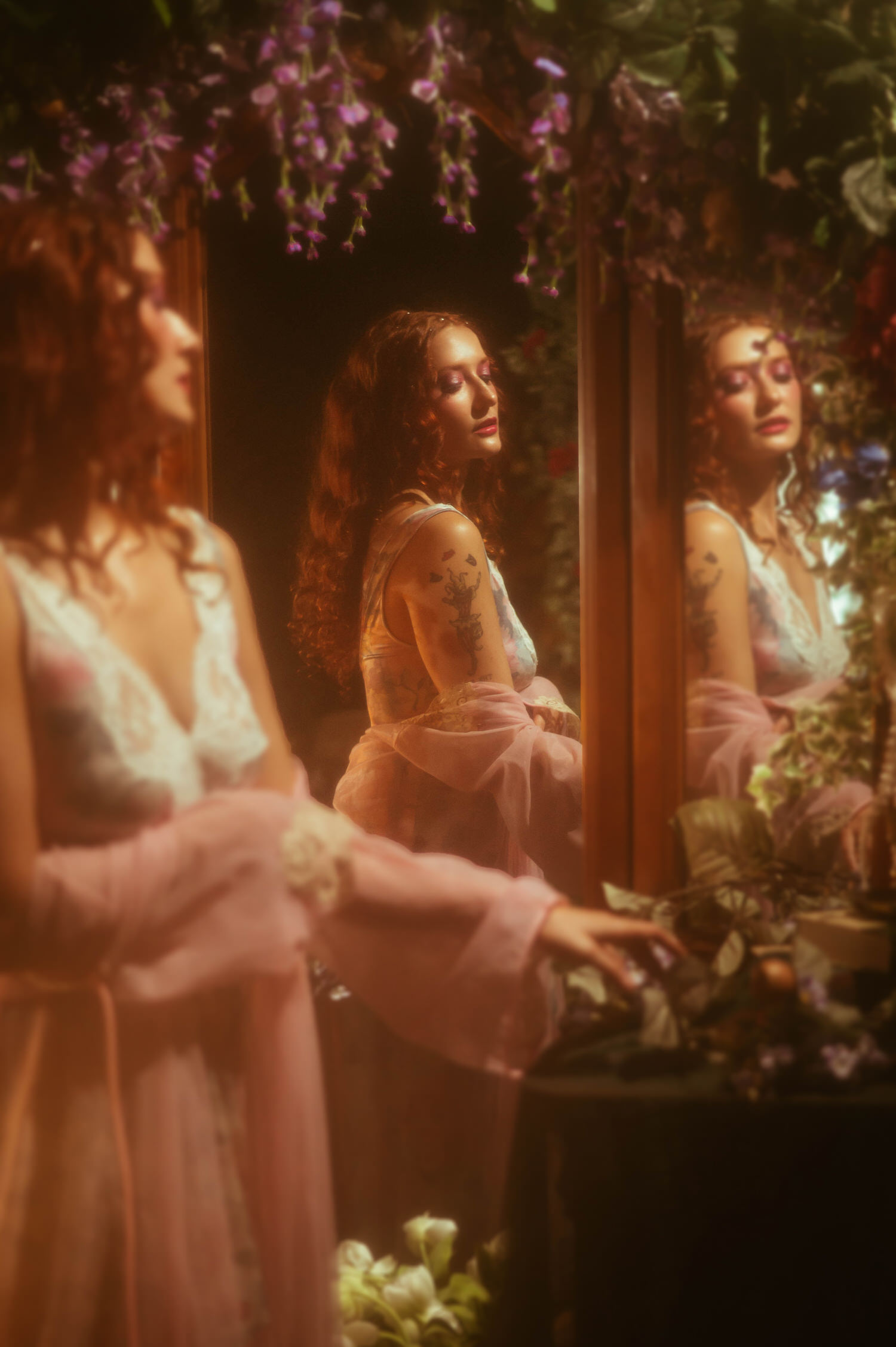 A woman in a vintage lace nightgown and flowing pink robe gazes downward near an antique mirror, framed by purple florals in this dreamy photography studio setting. The romantic styling reflects the magic of a Midsummer Night's Dream inspired session in the Dallas area. Royal Lune Photo.
