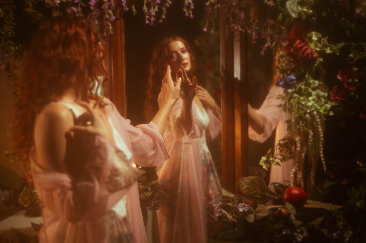 A model in a sheer pink robe and vintage nightgown reaches toward her mirrored reflection, surrounded by lush florals and warm candlelight in a fine art Midsummer Night's Dream photoshoot. This dreamy photography studio image was captured near Dallas. Royal Lune Photo.
