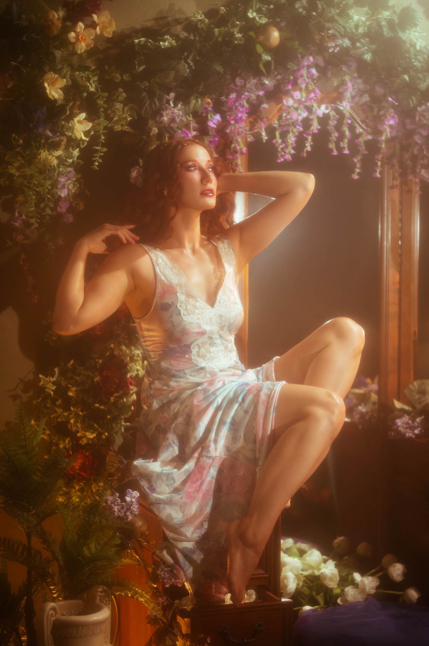 A woman in a white lace vintage nightgown poses beneath hanging wisteria and floral arrangements, showcasing dreamy photography style at a Dallas area studio. The warm golden light and lush greenery evoke the magic of A Midsummer Night's Dream. Royal Lune Photo.