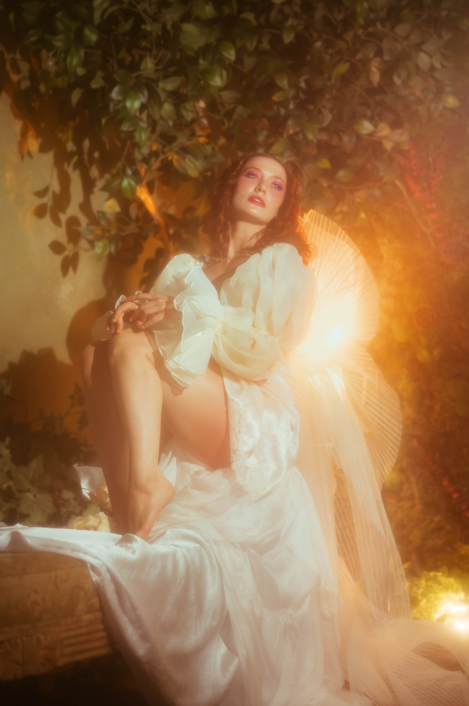 A model in a flowing white vintage nightgown with voluminous sleeves leans back in warm golden light beside an ornate fan, reflecting the fine art style of a Midsummer Night's Dream photoshoot. This dreamy photography studio image was captured near Dallas. Royal Lune Photo.