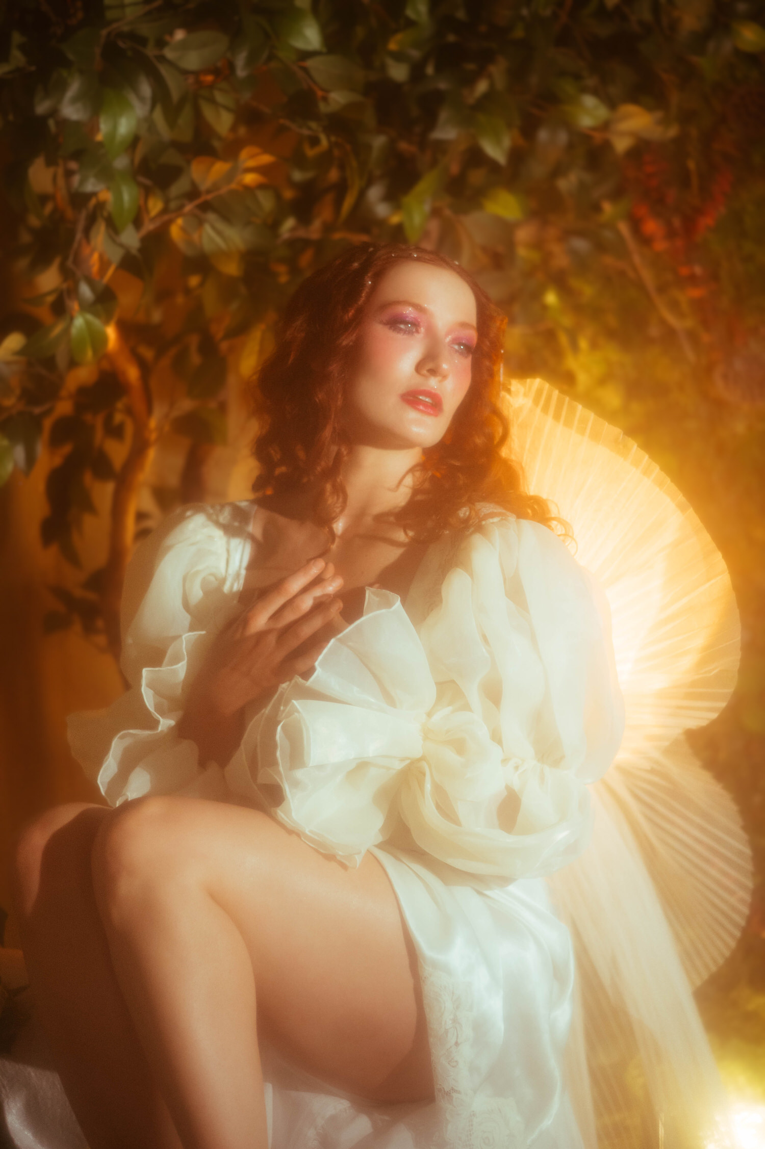 A model in a flowing white vintage nightgown with dramatic ruffled sleeves sits bathed in warm golden light beside an ornate fan, capturing the dreamy photography studio style of a Midsummer Night's Dream inspired session. This fine art portrait was created at a Dallas area studio. Royal Lune Photo.