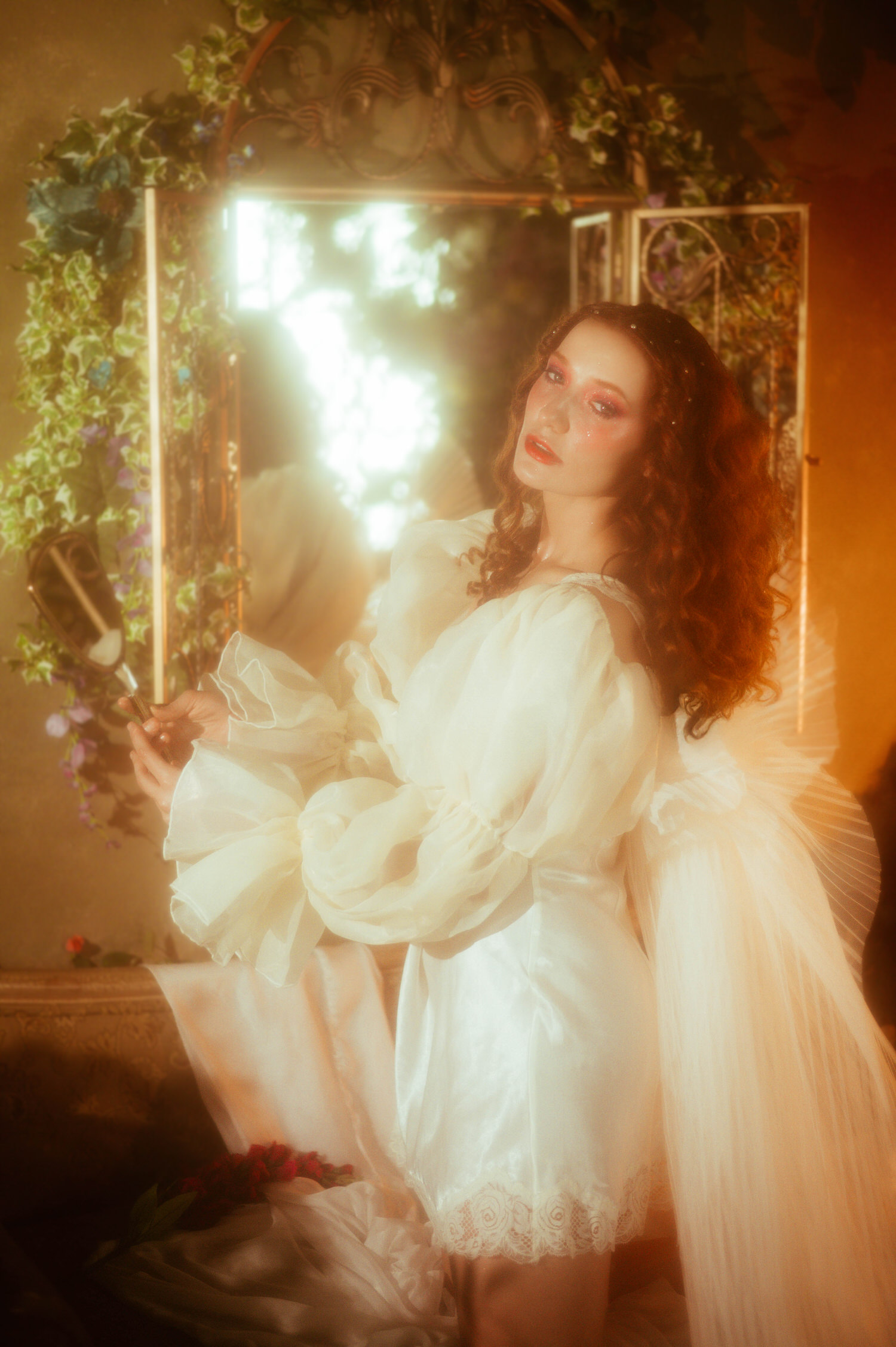 A model in a flowing white vintage nightgown with dramatic puffed sleeves holds an antique hand mirror amid wrought iron and trailing florals, capturing the dreamy photography style of a Midsummer Night's Dream inspired session. This fine art portrait was created at a Dallas area studio. Royal Lune Photo.