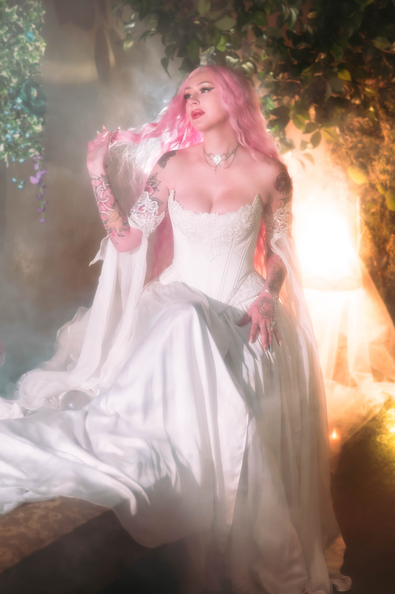 Fantasy bridal photos of bride seated in a lace corset fantasy bridal gown with flowing chiffon sleeves, pink hair glowing in soft haze at Royal Lune Studio.