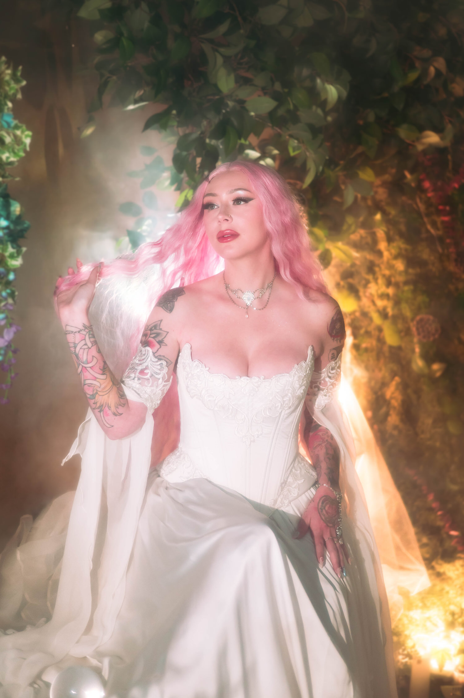 Close portrait of bride in a fairytale bridal gown, lifting her pink hair beneath hanging greenery in a romantic fantasy wedding photography setting.