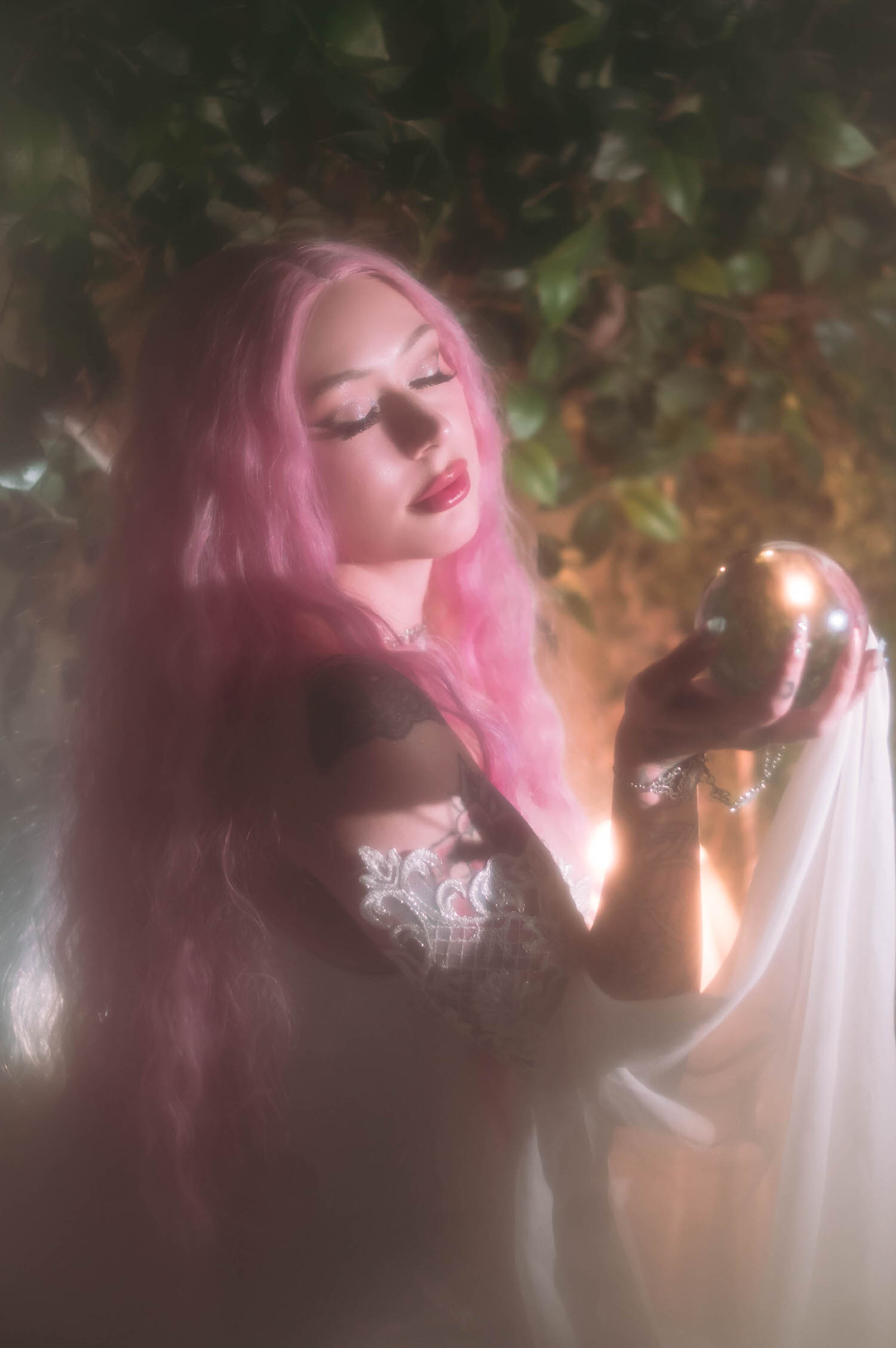 Dreamy close-up of bride in a fantasy wedding photography scene, eyes closed while holding a glass orb against a glowing greenery backdrop.