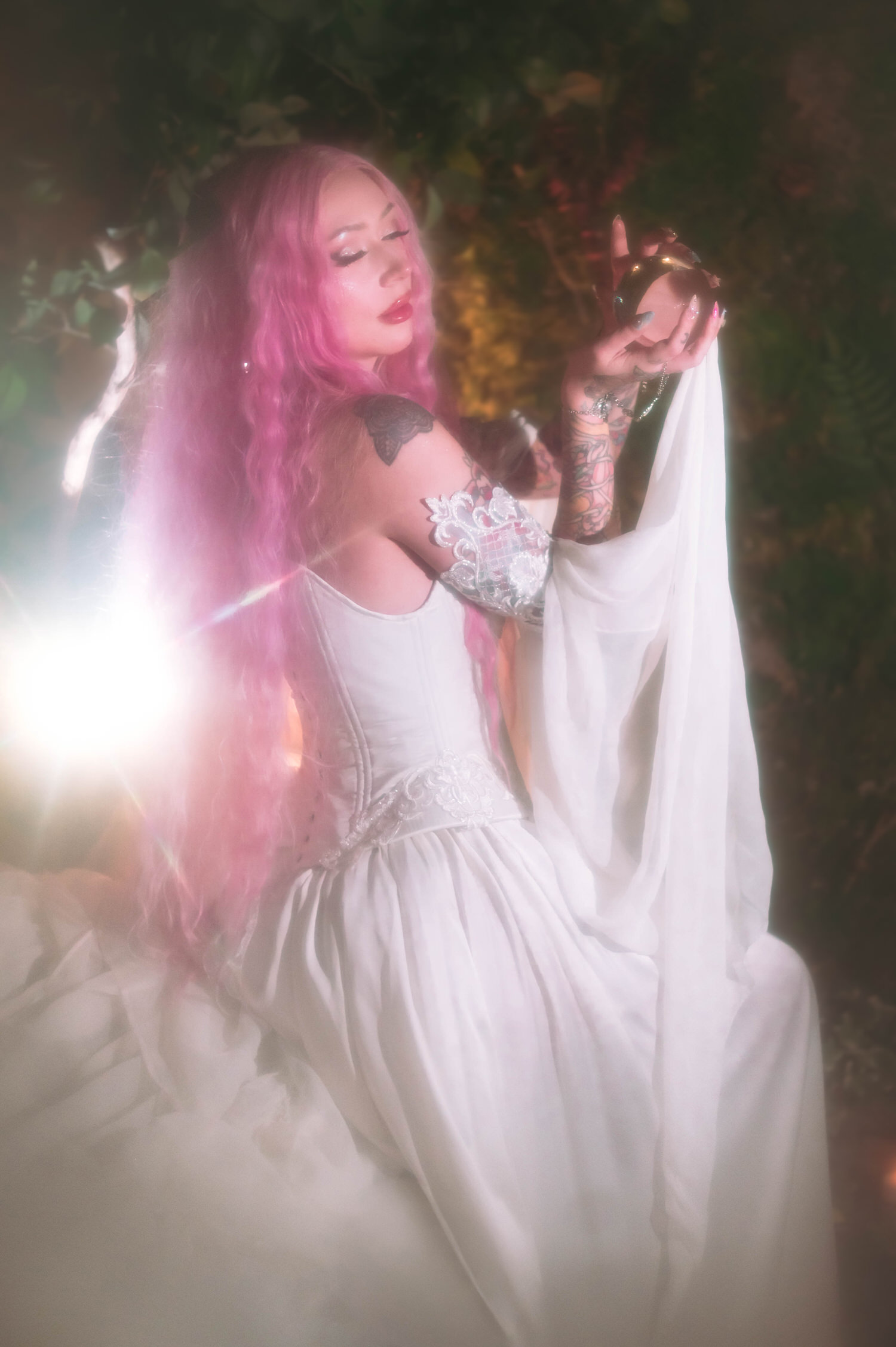 Romantic side portrait of bride holding a glass orb in a firefly path bridal gown, illuminated by soft glowing backlight and lush greenery.
