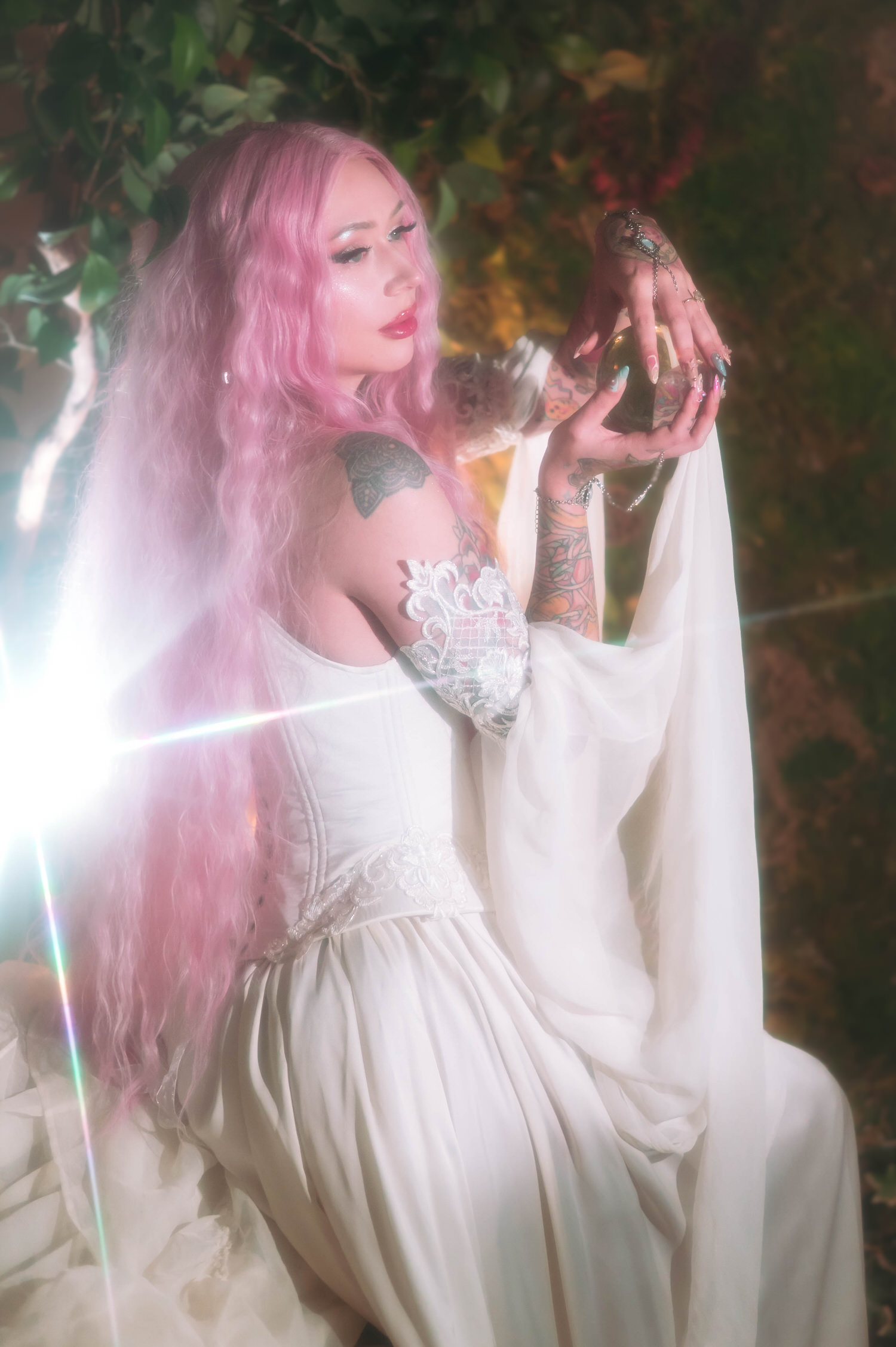 Fantasy bridal photos of bride with long pink hair holding a golden orb, wearing a lace corset fantasy bridal gown with flowing chiffon sleeves against a lush greenery backdrop at Royal Lune Studio.
