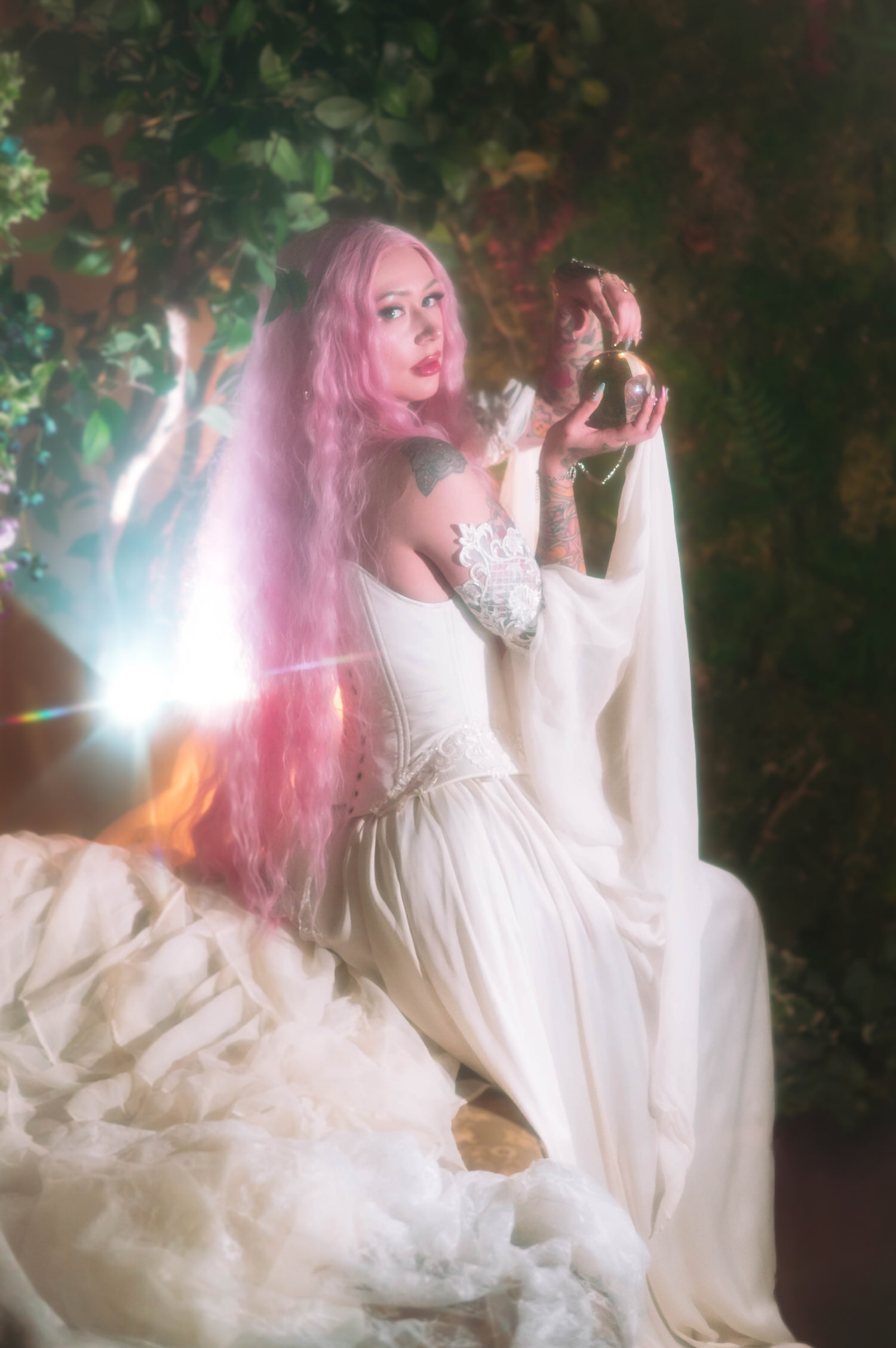Fantasy bridal photos of a bride with long pink hair seated in a flowing ivory fantasy bridal gown, holding a golden orb in a lush greenery backdrop at Royal Lune Studio.