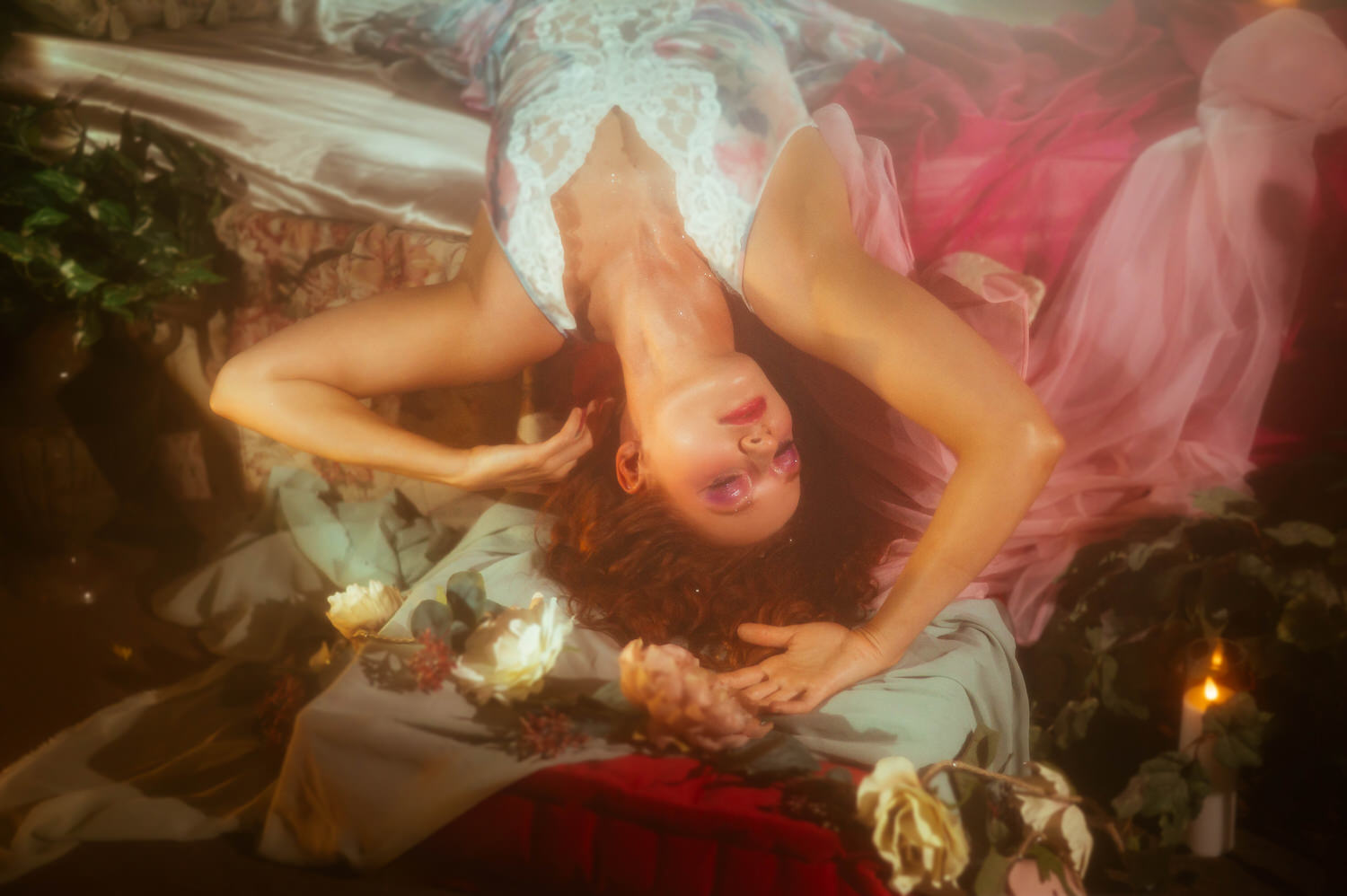 A model in a lace vintage nightgown and pink robe lies among florals and candlelight, captured from an artistic angle for a Midsummer Night's Dream inspired session. This dreamy photography studio portrait was created at a Dallas area studio. Royal Lune Photo.