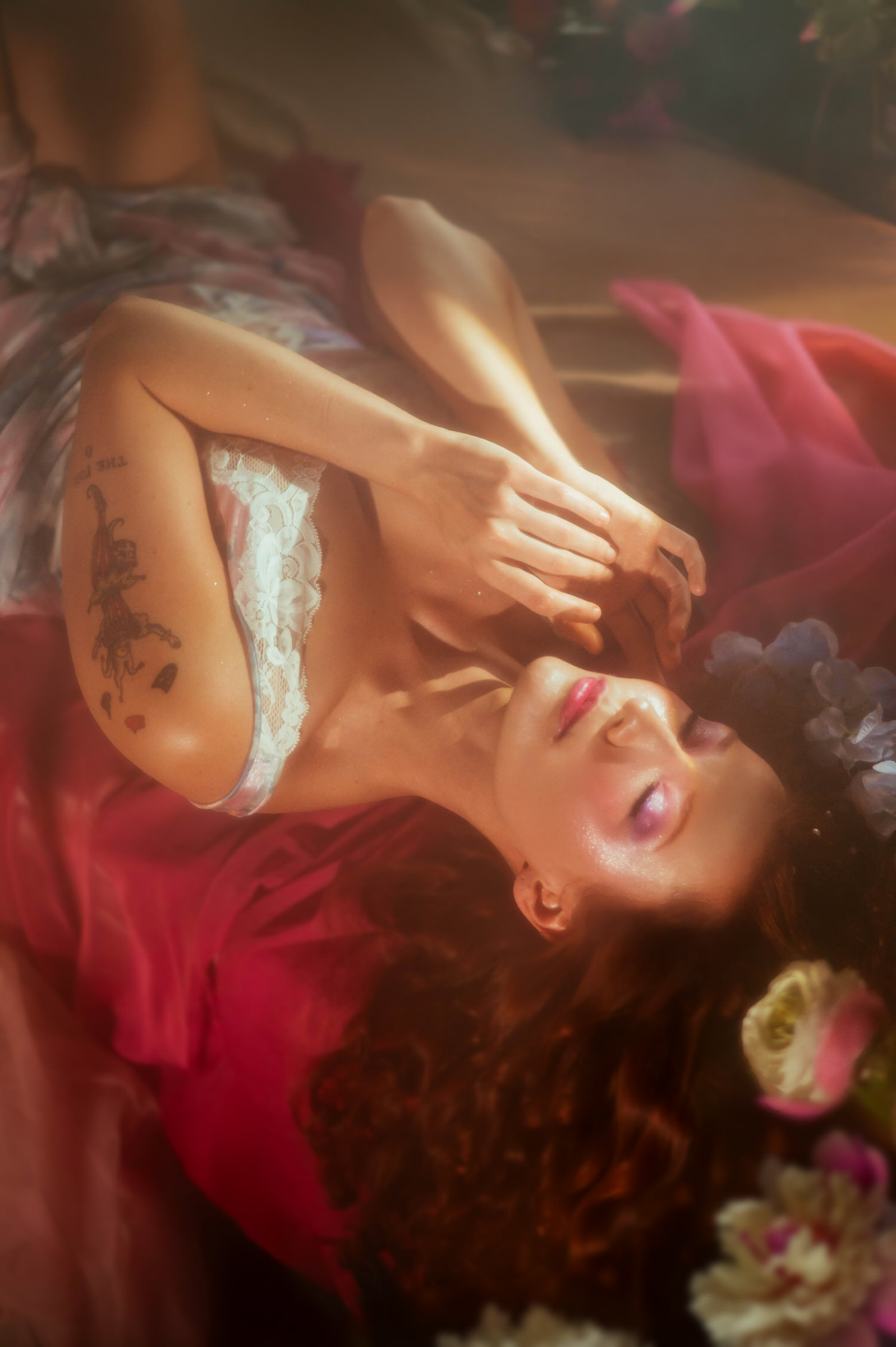 A model in vintage lace lingerie and a deep red wrap leans back among florals, captured from above for a fine art Midsummer Night's Dream inspired session. This dreamy photography studio image highlights soft golden lighting near Dallas. Royal Lune Photo.