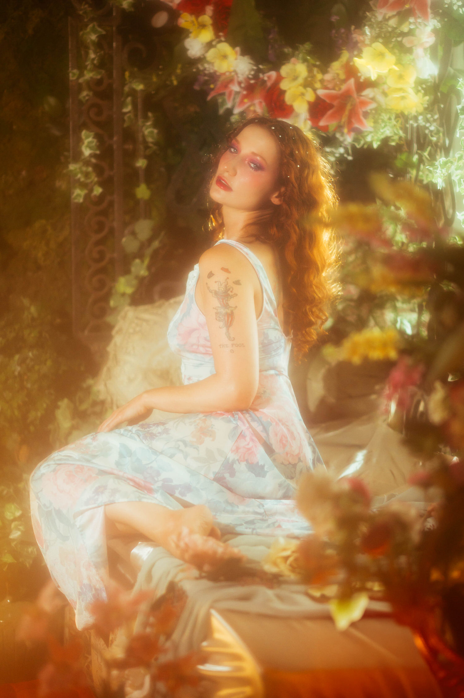 A red haired model in a floral vintage nightgown glances over her shoulder amid warm golden light and blooming flowers, reflecting the dreamy photography studio aesthetic of a Midsummer Night's Dream shoot. This fine art portrait was captured near Dallas. Royal Lune Photo.