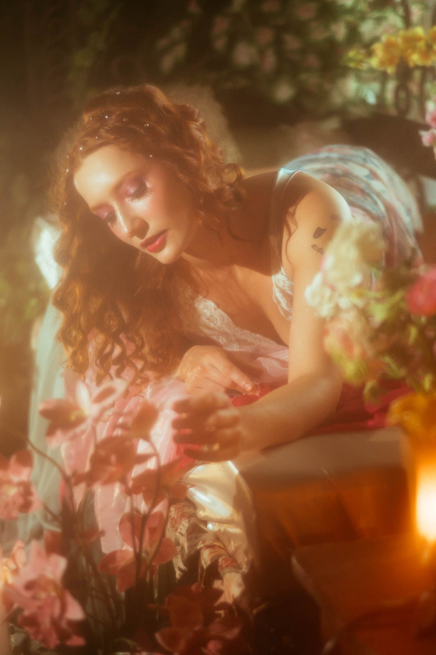 A model in a vintage lace nightgown leans forward among soft golden light and pink florals, captured in a dreamy photography style for a Midsummer Night's Dream photoshoot. This fine art session was shot at a Dallas area studio. Royal Lune Photo.