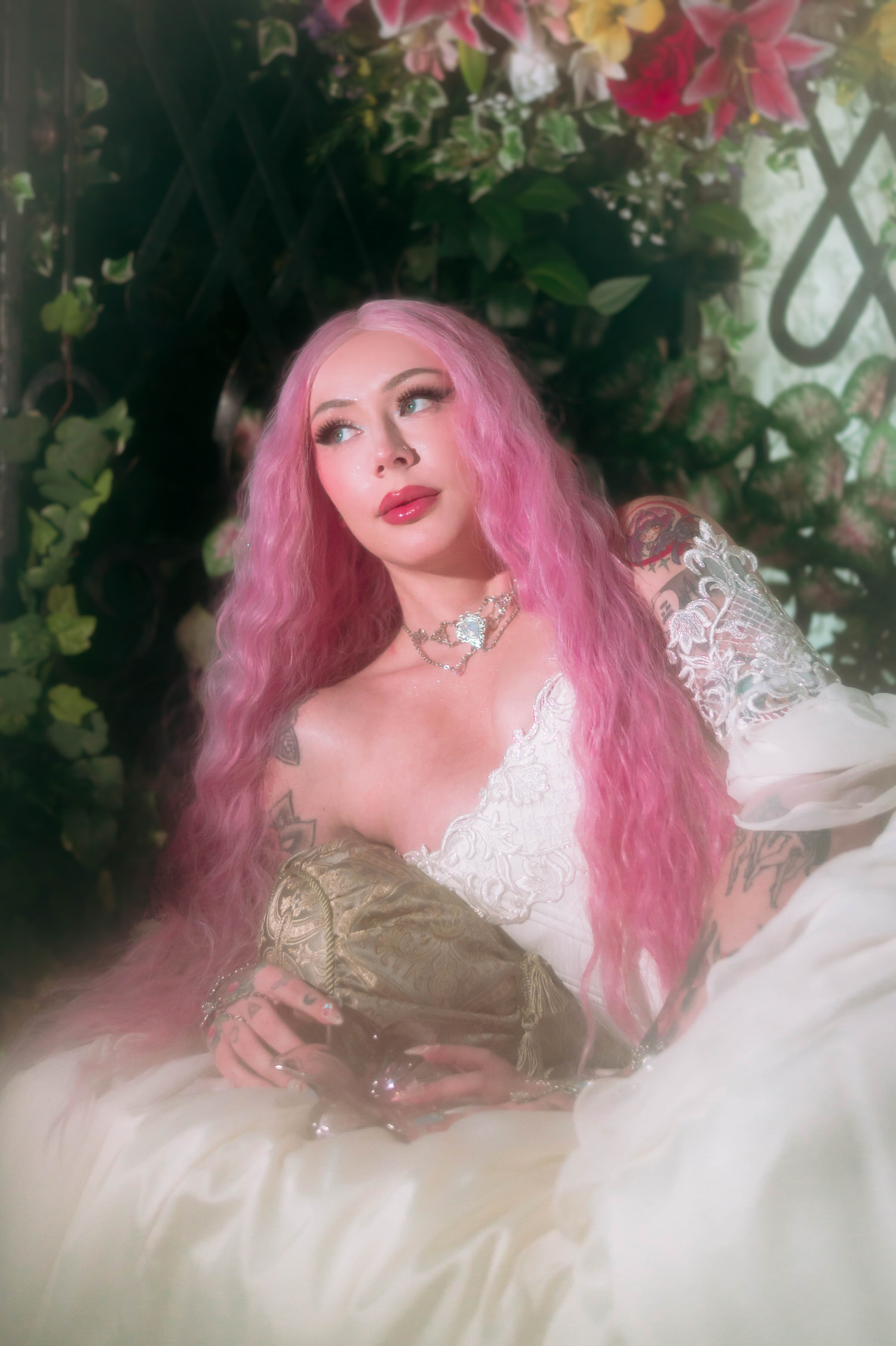 Reclining bride in a bridal gown by firefly path, surrounded by pink lilies and ivy against an ornate wrought iron backdrop.