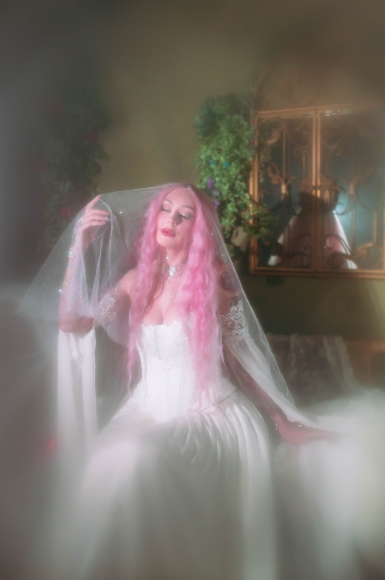 Bride seated on vintage sofa in a fairytale bridal gown with sparkling veil, styled for fantasy wedding photography with ivy and ornate gold window frame.