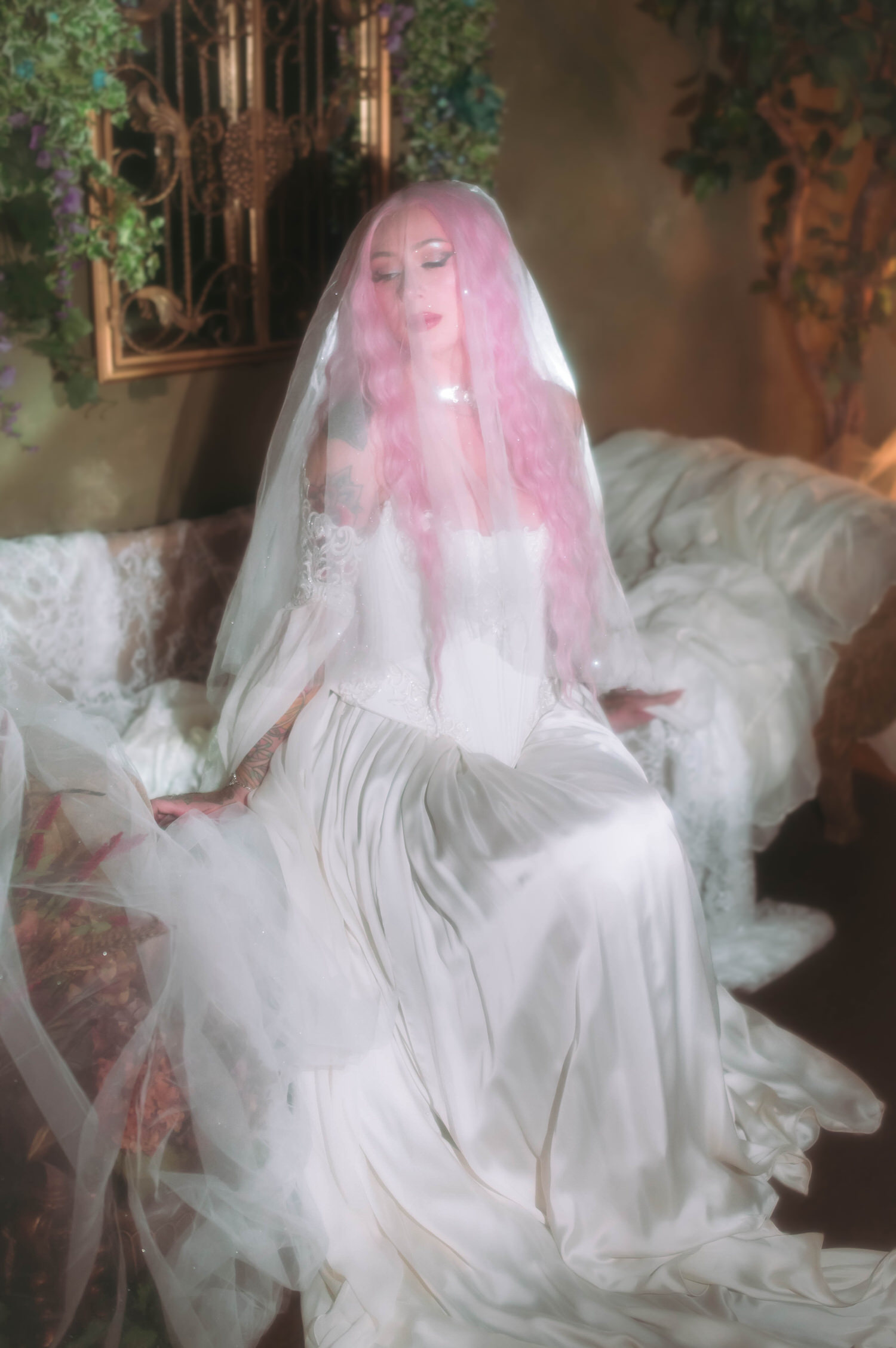 Fantasy bridal photos of bride sitting gracefully in a satin and lace fantasy bridal gown, veil draped over her hair with romantic haze and greenery.