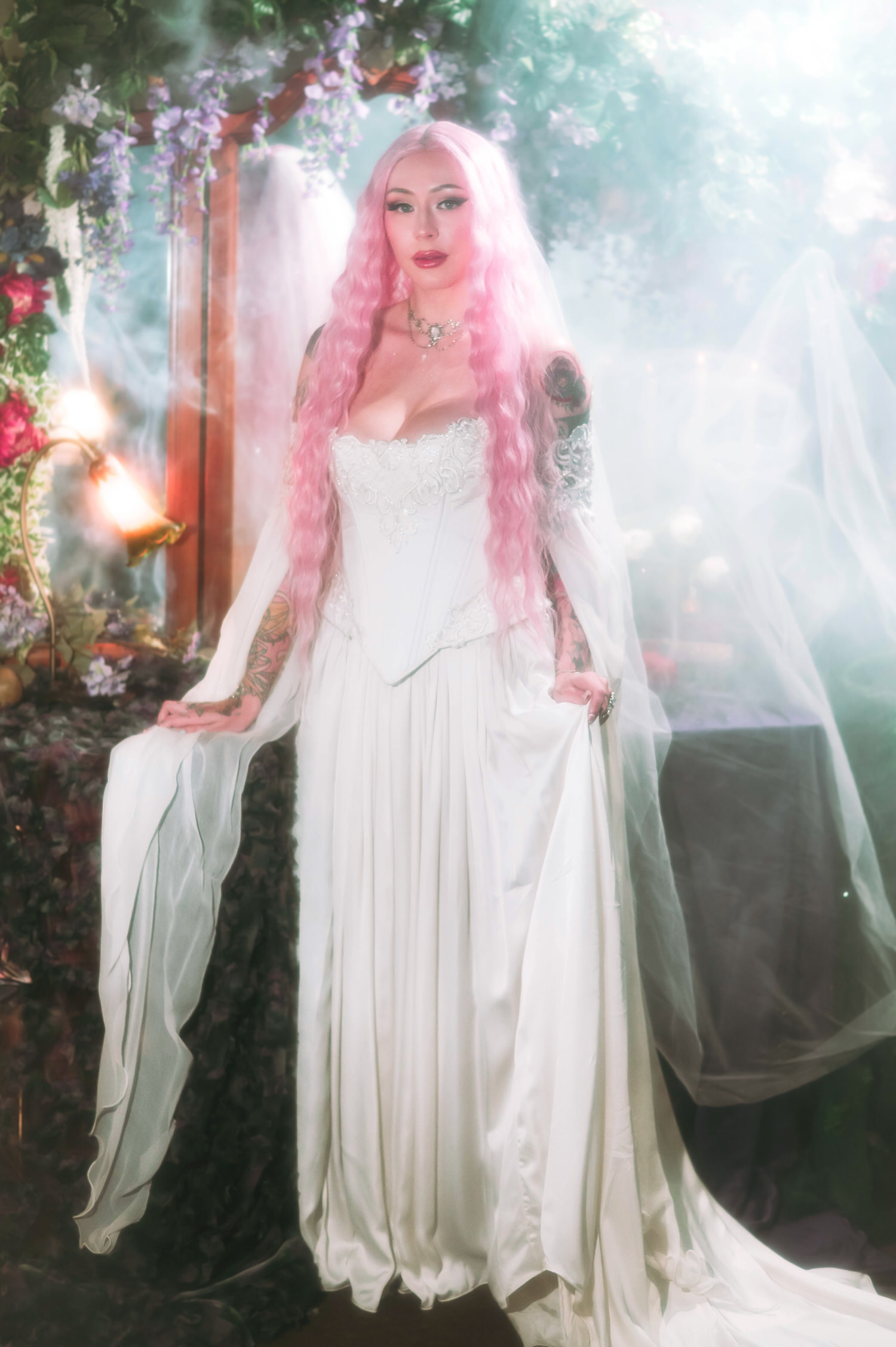 Full-length fantasy bridal photos of bride standing in a lace corset fantasy bridal gown with flowing chiffon sleeves, pink hair cascading beneath a veil in a misty fairytale wedding setting.