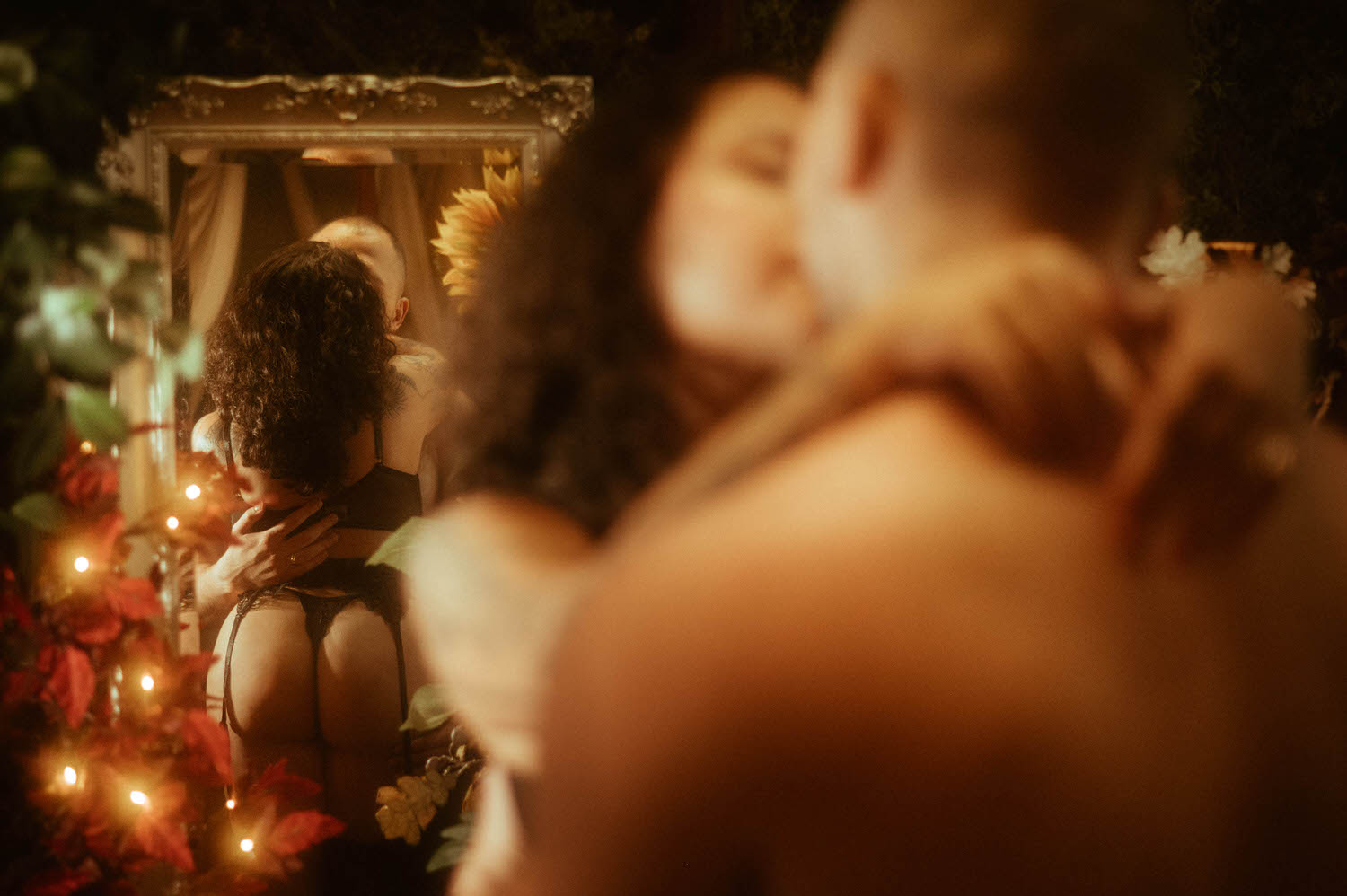 A couple embraces in front of an ornate gold-framed mirror surrounded by warm string lights, red poinsettias, a sunflower, and lush botanicals, with the woman's black lingerie and garter belt reflected in the glass in this couples boudoir guide to intimate fine art photography at Royal Lune Photo.