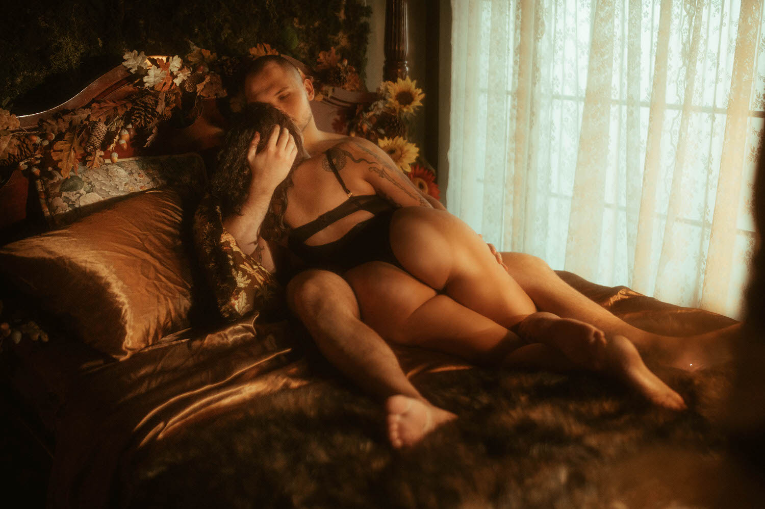 A couple reclines together on a bronze satin bed surrounded by autumn sunflowers, oak leaves, and pine cones with soft lace curtain light streaming in from the window, captured during a Dallas couples boudoir session at Royal Lune Photo.