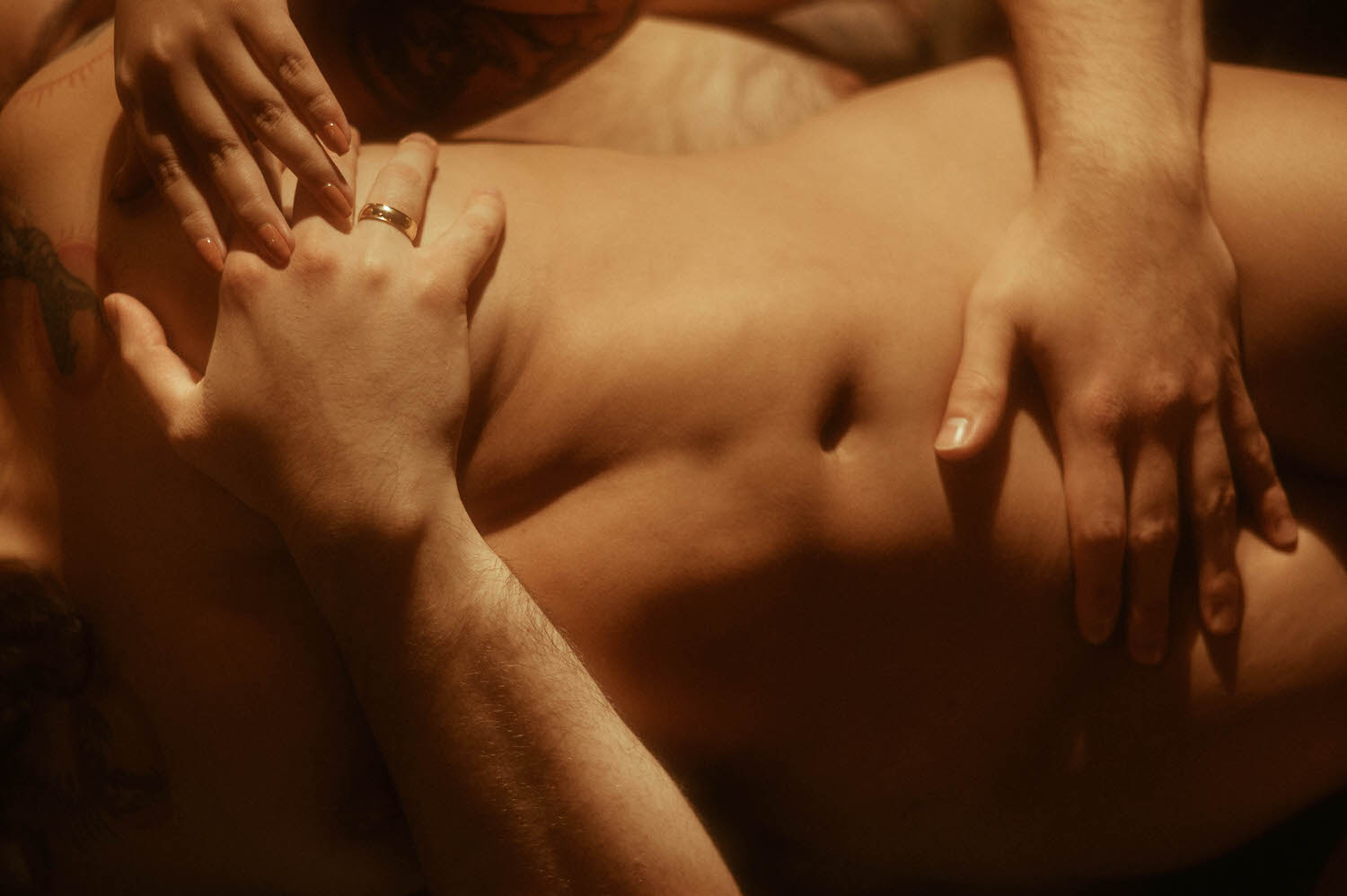 An intimate detail shot of two pairs of hands resting on a woman's bare torso bathed in deep amber light, with a gold wedding band catching the glow, showcasing couples boudoir posing tips centered on the quiet power of touch at Royal Lune Photo.